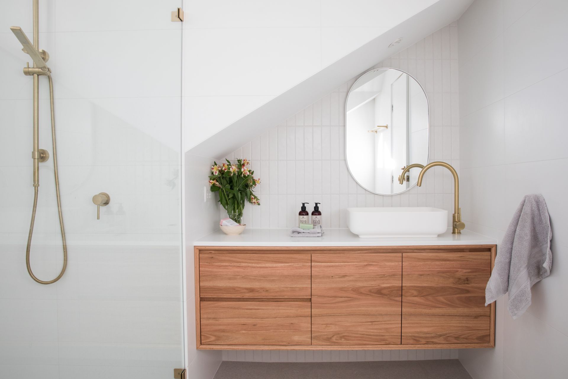 Bright modern bathroom vanity with wood cabinet, round mirror, white sink, gold faucet, and glass shower