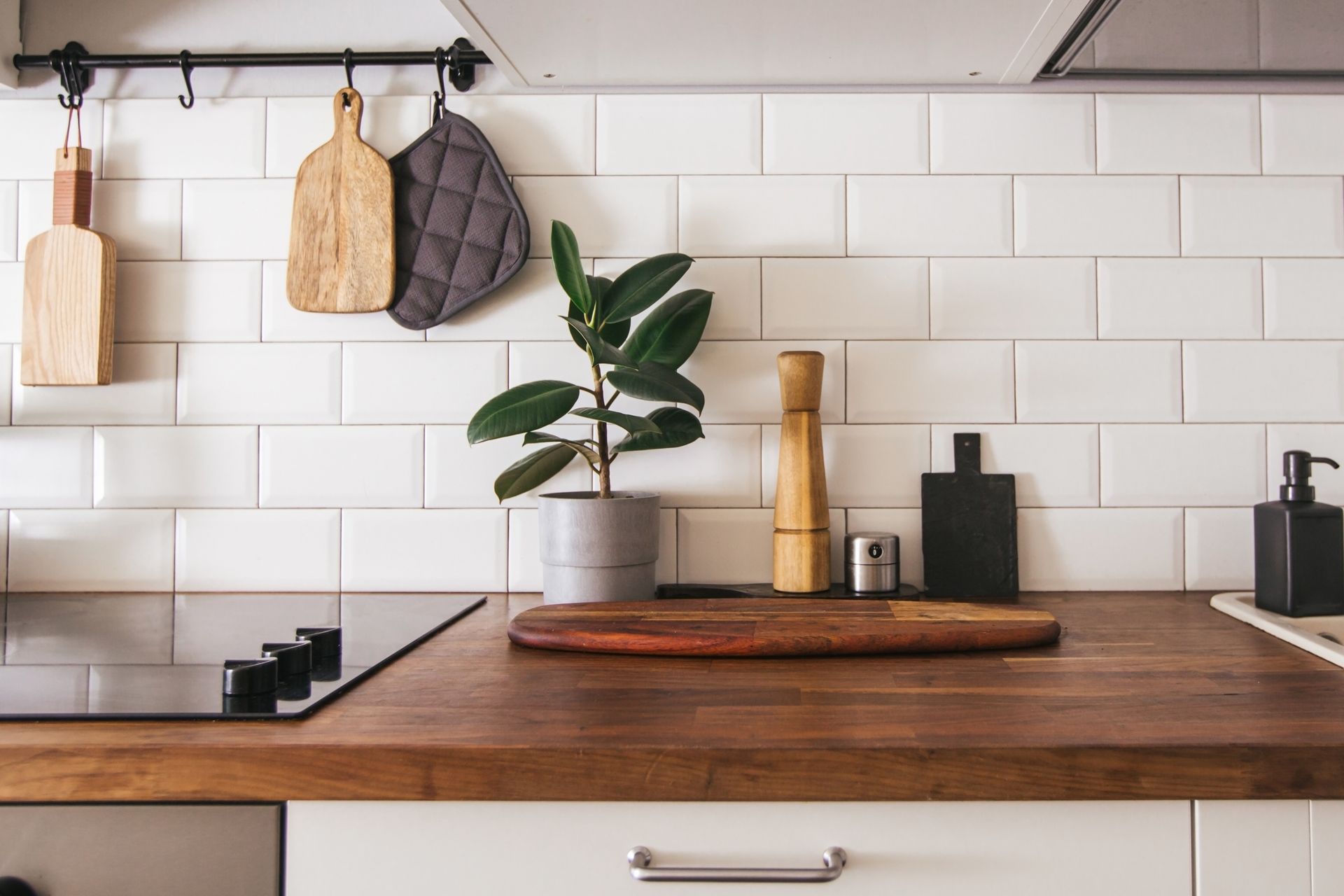 Modern kitchen countertop with wooden cutting board, potted plant, utensils, and hanging towels on white tile backsplash Modern kitchen countertop with wooden cutting board, potted plant, utensils, and hanging towels on white tile backsplash