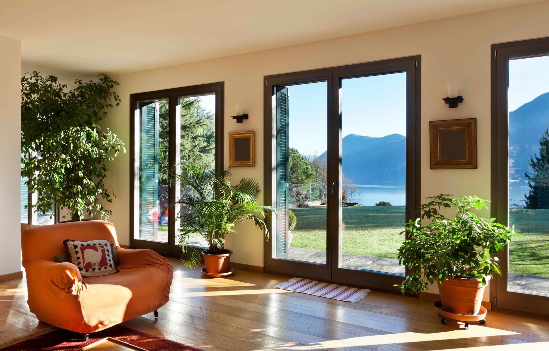 Sunlit living room with orange chair, potted plants, and large windows overlooking a lake and mountains Sunlit living room with orange chair, potted plants, and large windows overlooking a lake and mountains