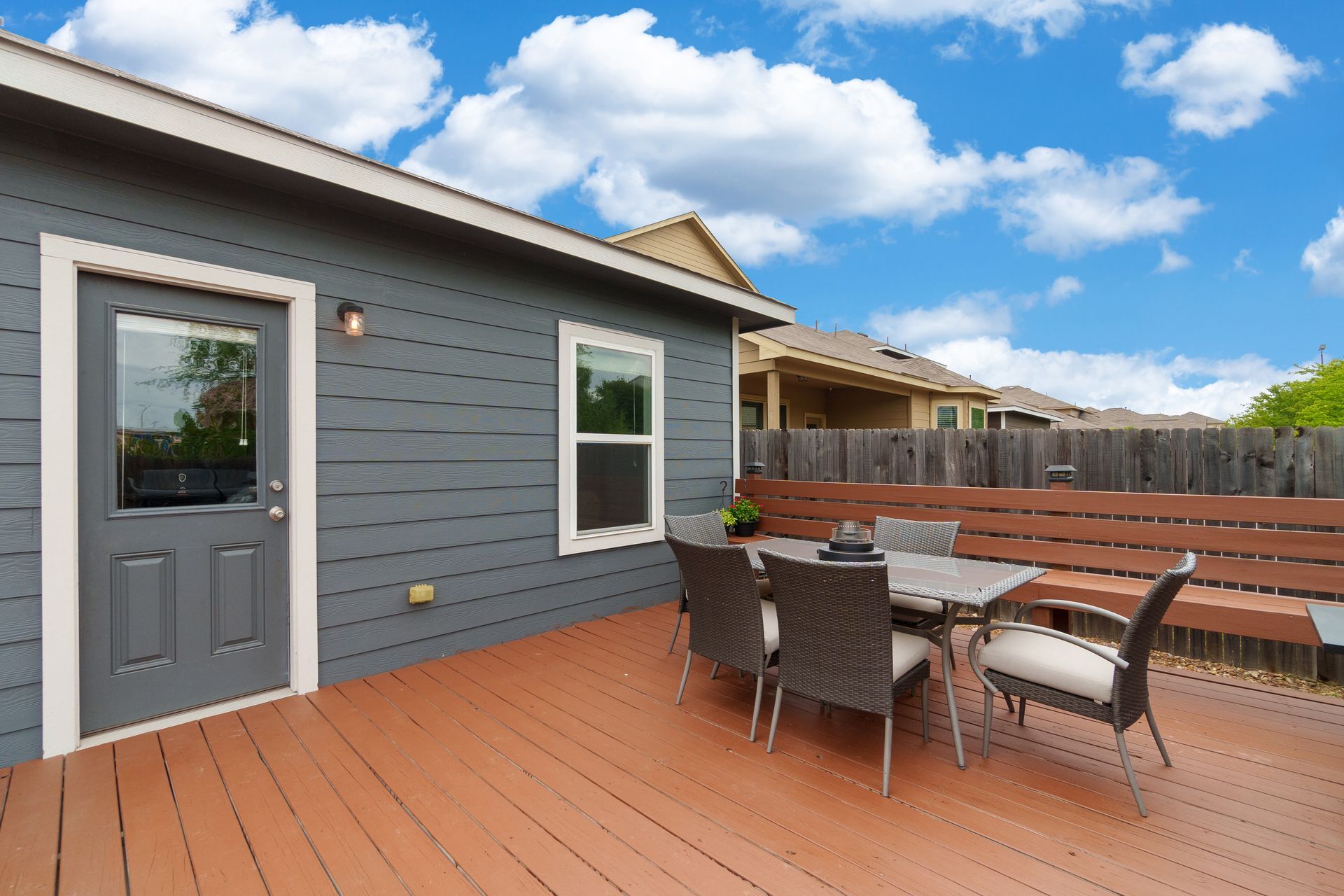 Outdoor wooden deck with patio table and chairs beside a gray house under a blue sky Outdoor wooden deck with patio table and chairs beside a gray house under a blue sky