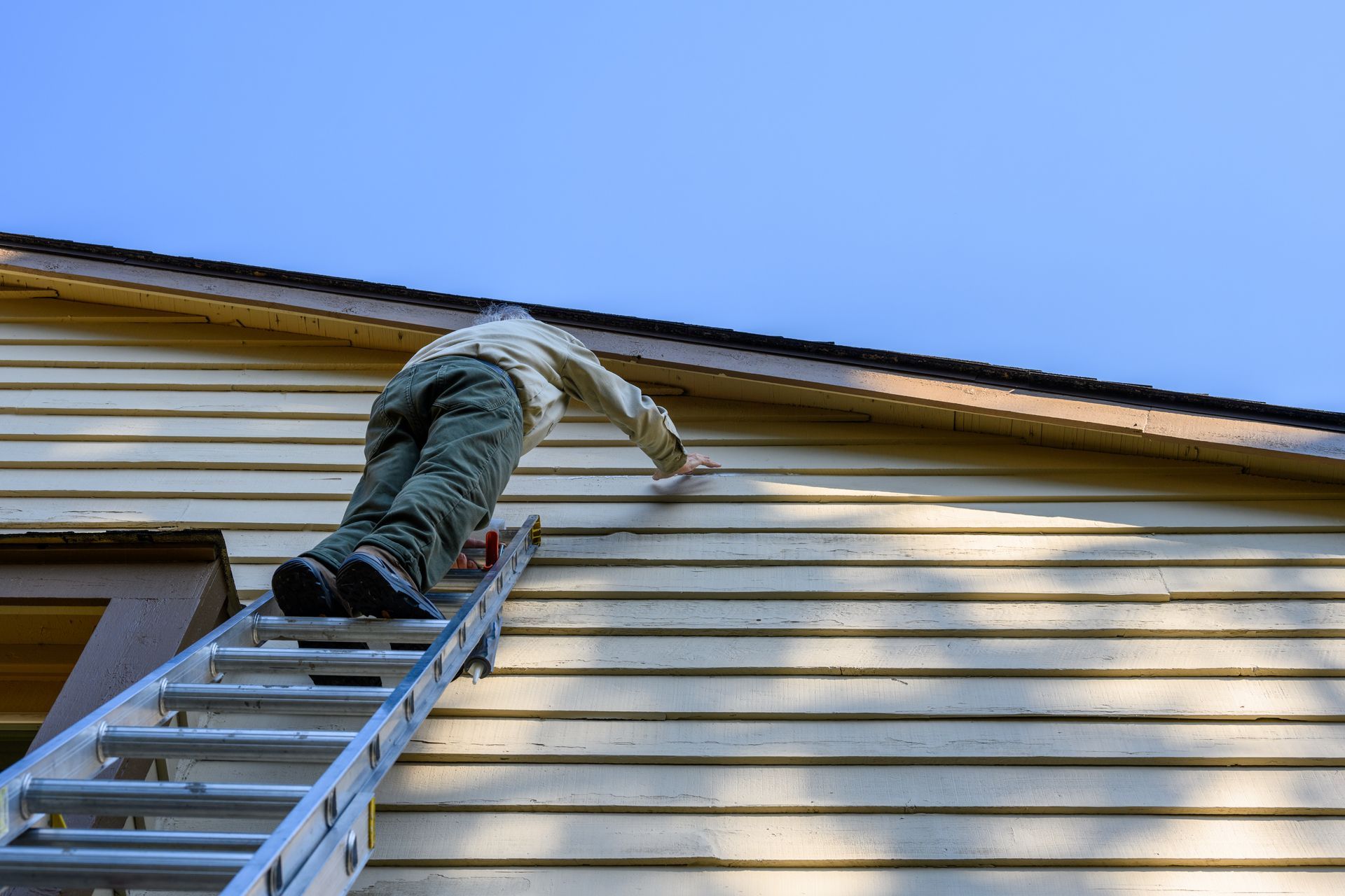 Worker on a ladder painting a house’s exterior siding under a blue sky