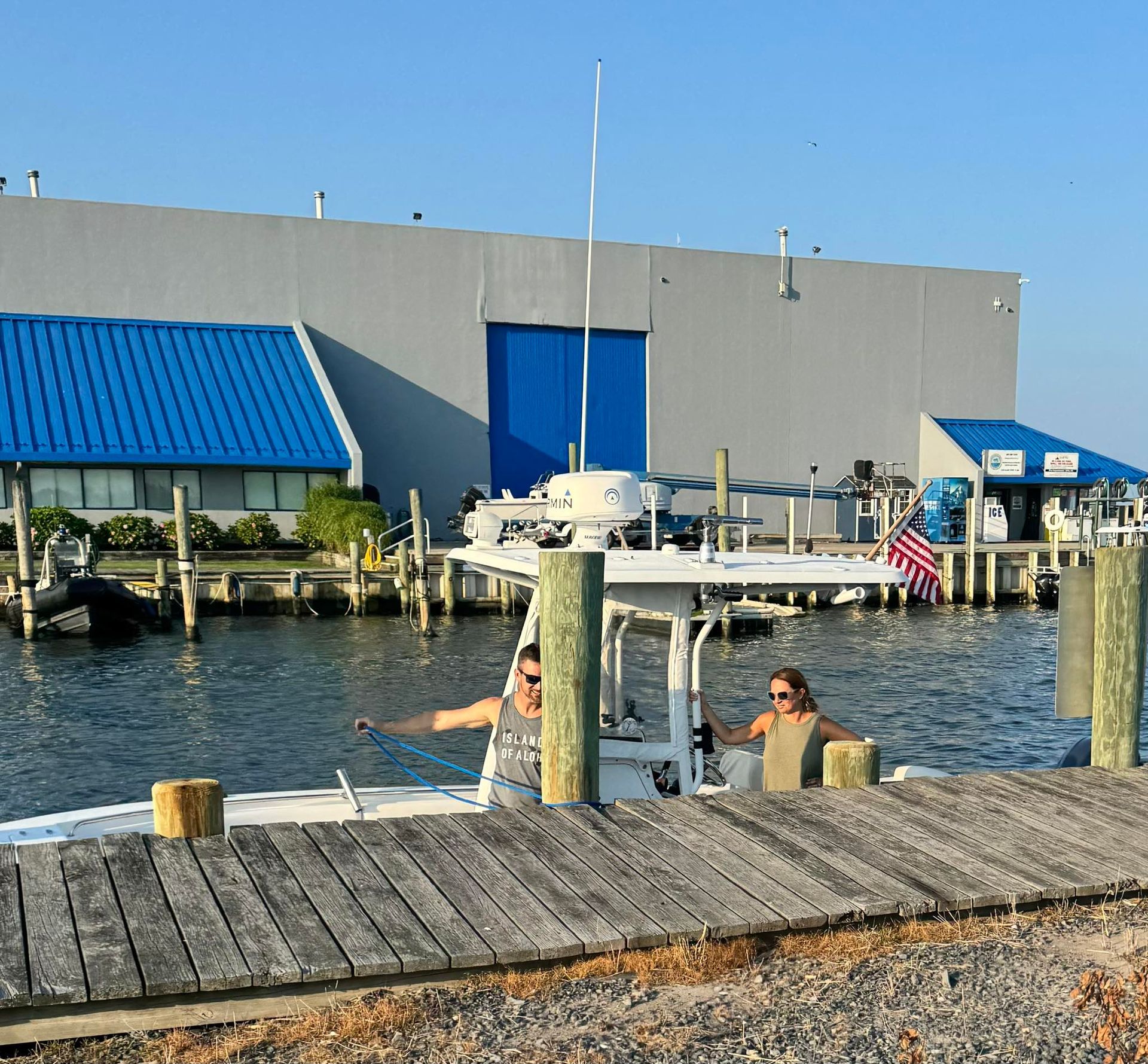 Harbor scene with a gray building, blue awnings, docked boats, and two people by the water under a clear sky