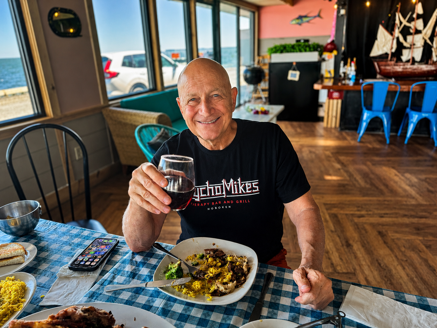 Smiling man at a seaside restaurant raises a drink over a checkered table with seafood plates
