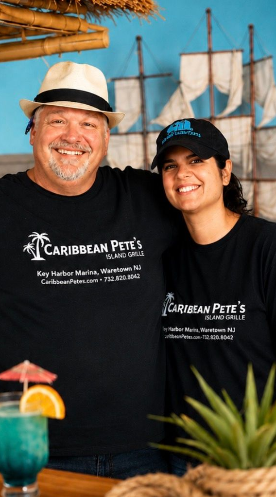 Two smiling staff members in black shirts and hats stand in front of a Caribbean Pirates sailboat backdrop.