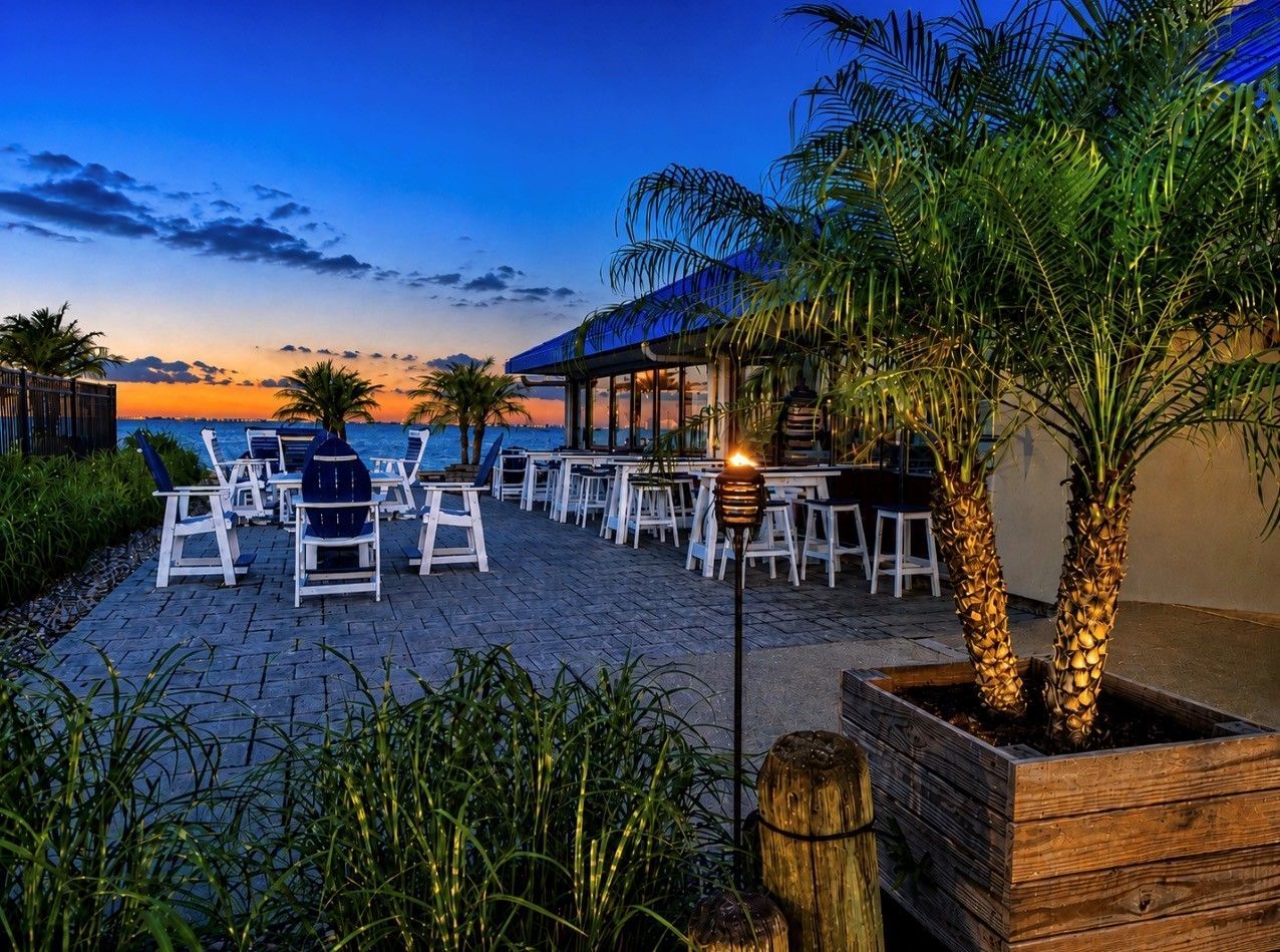 Beachfront patio at sunset with white chairs, tables, palm trees, and warm lights under a blue-orange sky