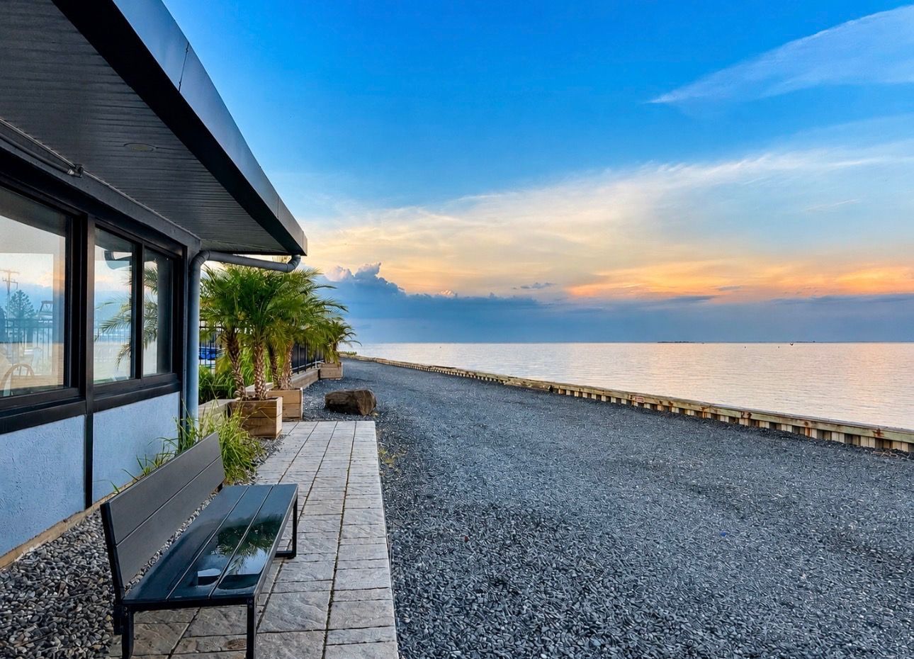Seaside patio beside a building, with bench seating and a gravel path overlooking calm water at sunset.