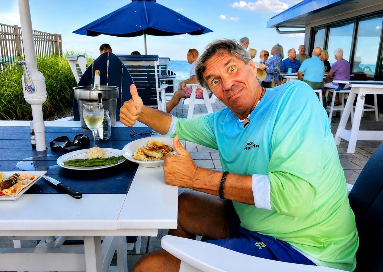 Man in a turquoise shirt giving thumbs up at an outdoor restaurant table with food and blue umbrellas.
