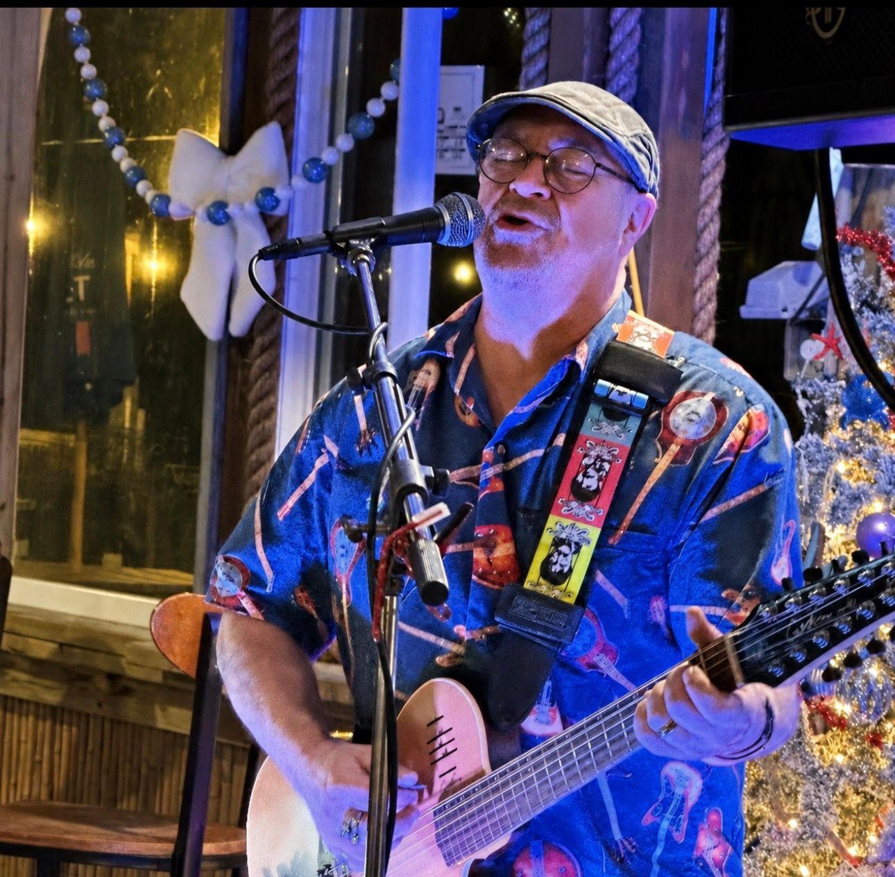 Singer-guitarist performing onstage under blue lights, wearing a colorful shirt and cap, singing into a microphone