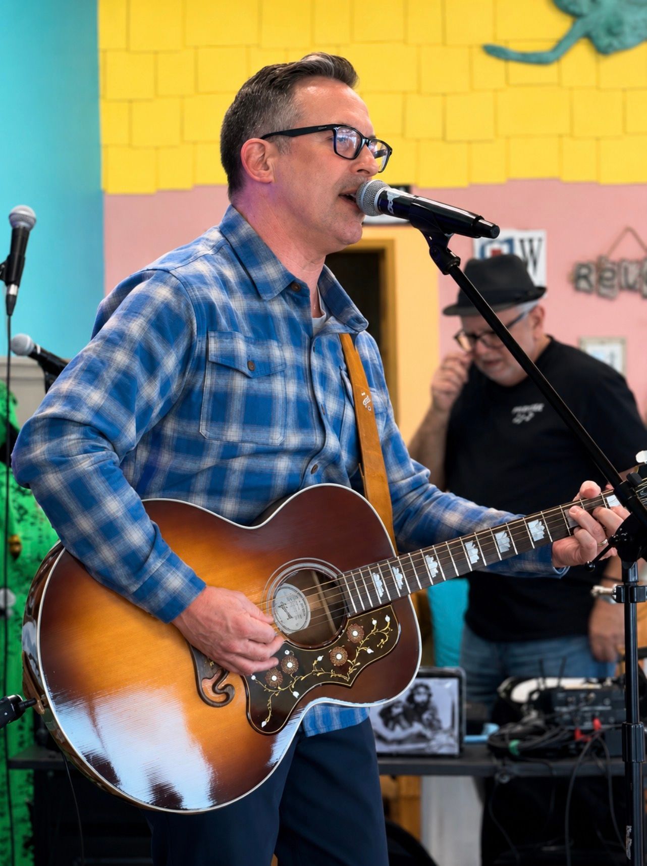 Man singing and playing acoustic guitar onstage in a blue plaid shirt