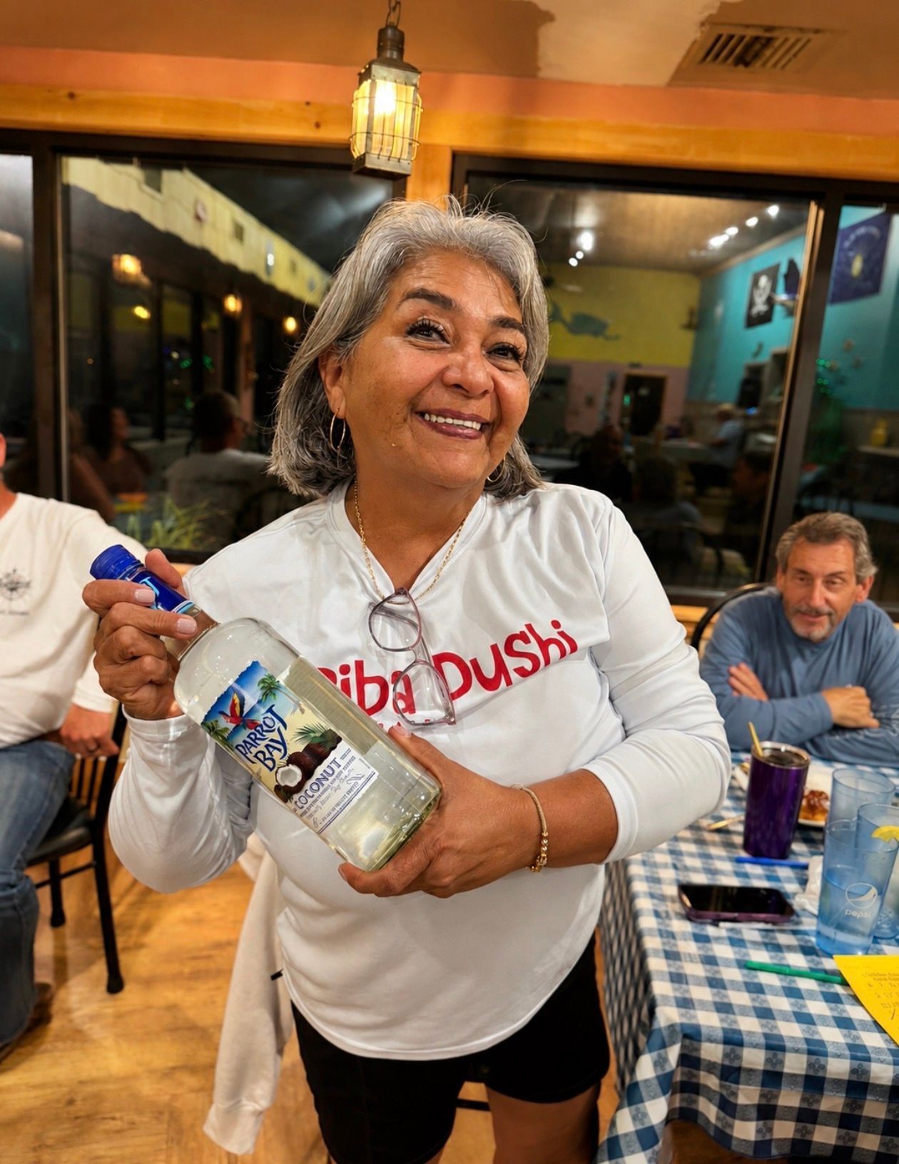 Smiling woman in a white shirt holding a bottle at a restaurant table with checkered tablecloths