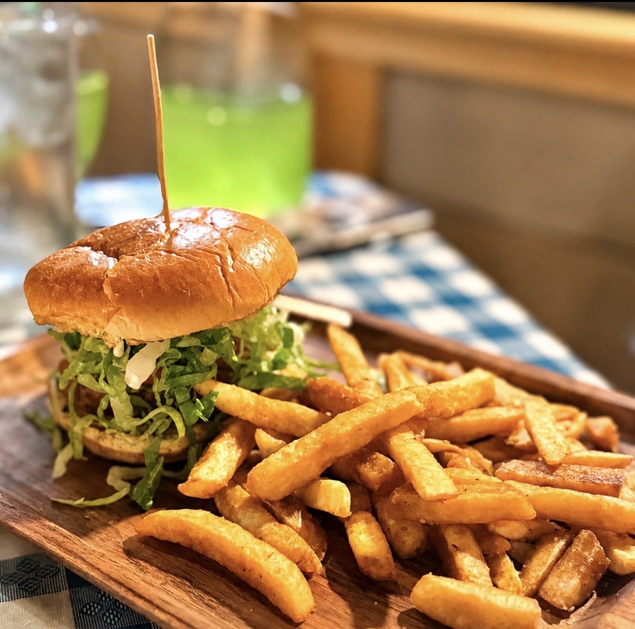Sandwich with fries on a wooden board, with a green drink and blue checkered tablecloth in the background