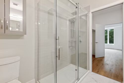 Bathroom with glass shower, toilet, and open doorway to a bedroom with hardwood floors.