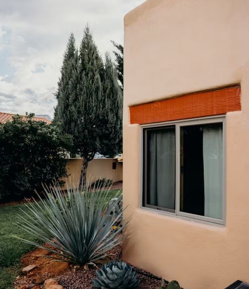 Tan stucco building with a window framed in brown. Tall green trees and blue agave plants in the yard.