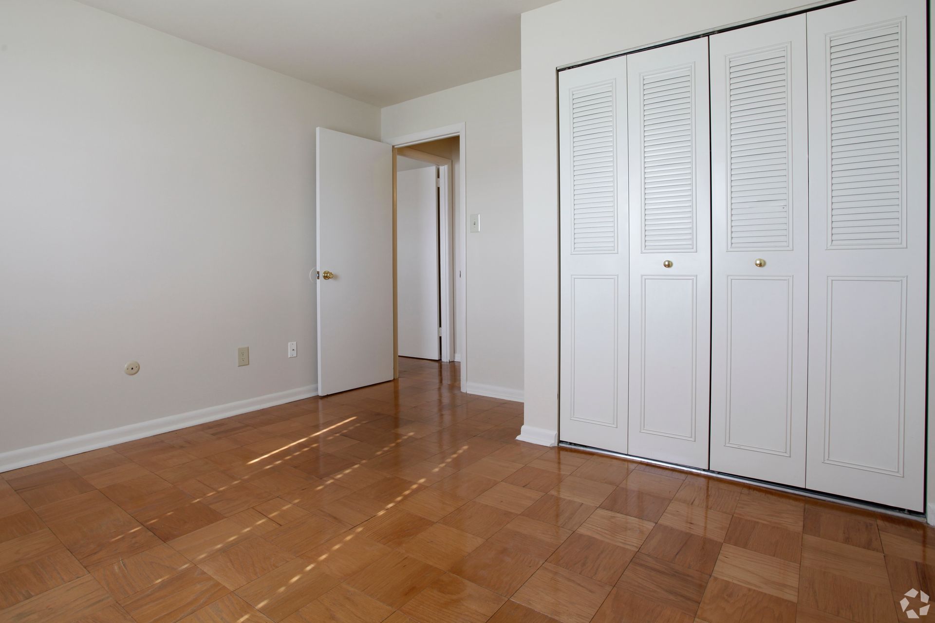 Empty room with light wood floors, white walls, closet doors, and open door to hallway.