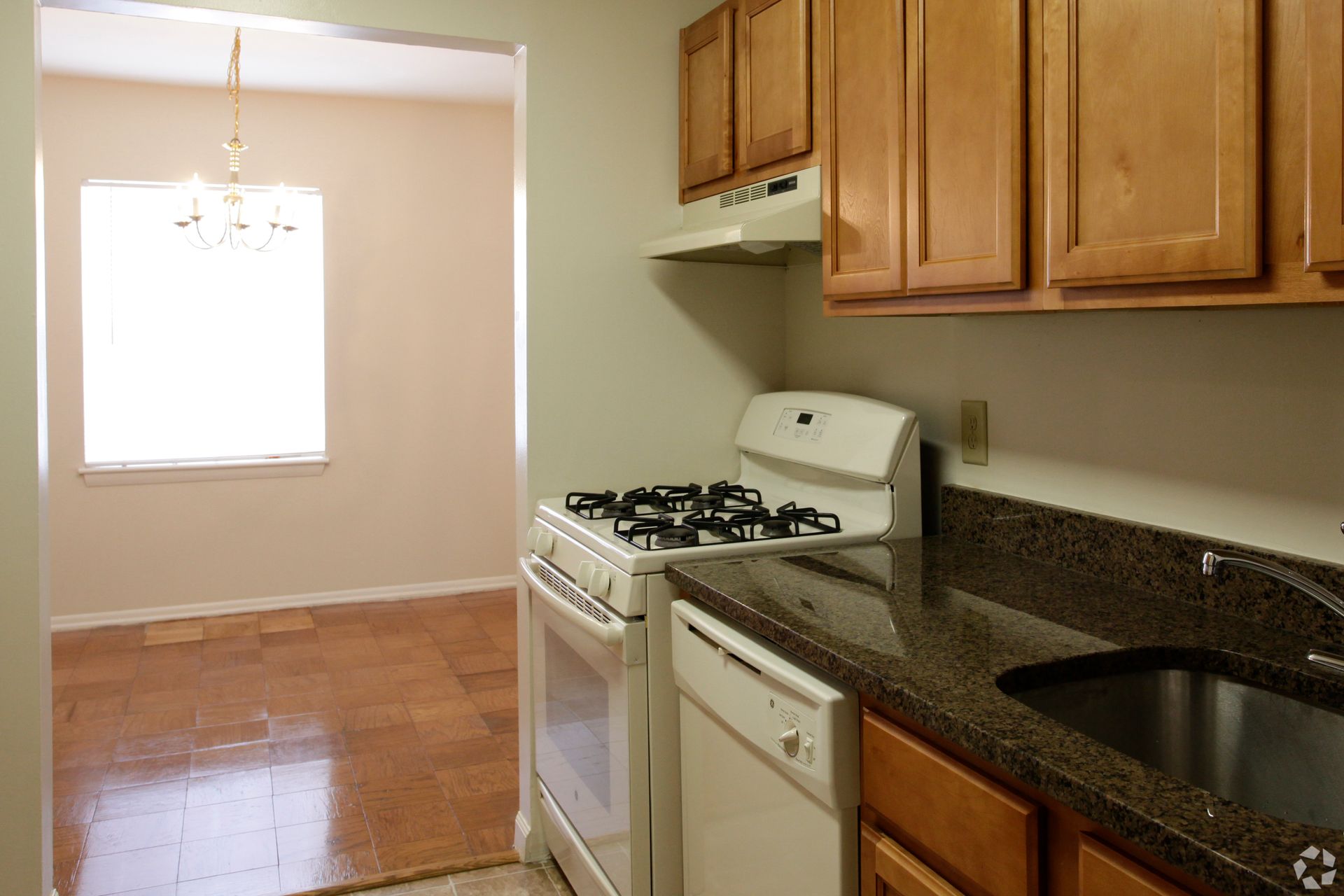 Kitchen with light brown cabinets, white appliances, and a dark countertop. Dining room visible in the background.