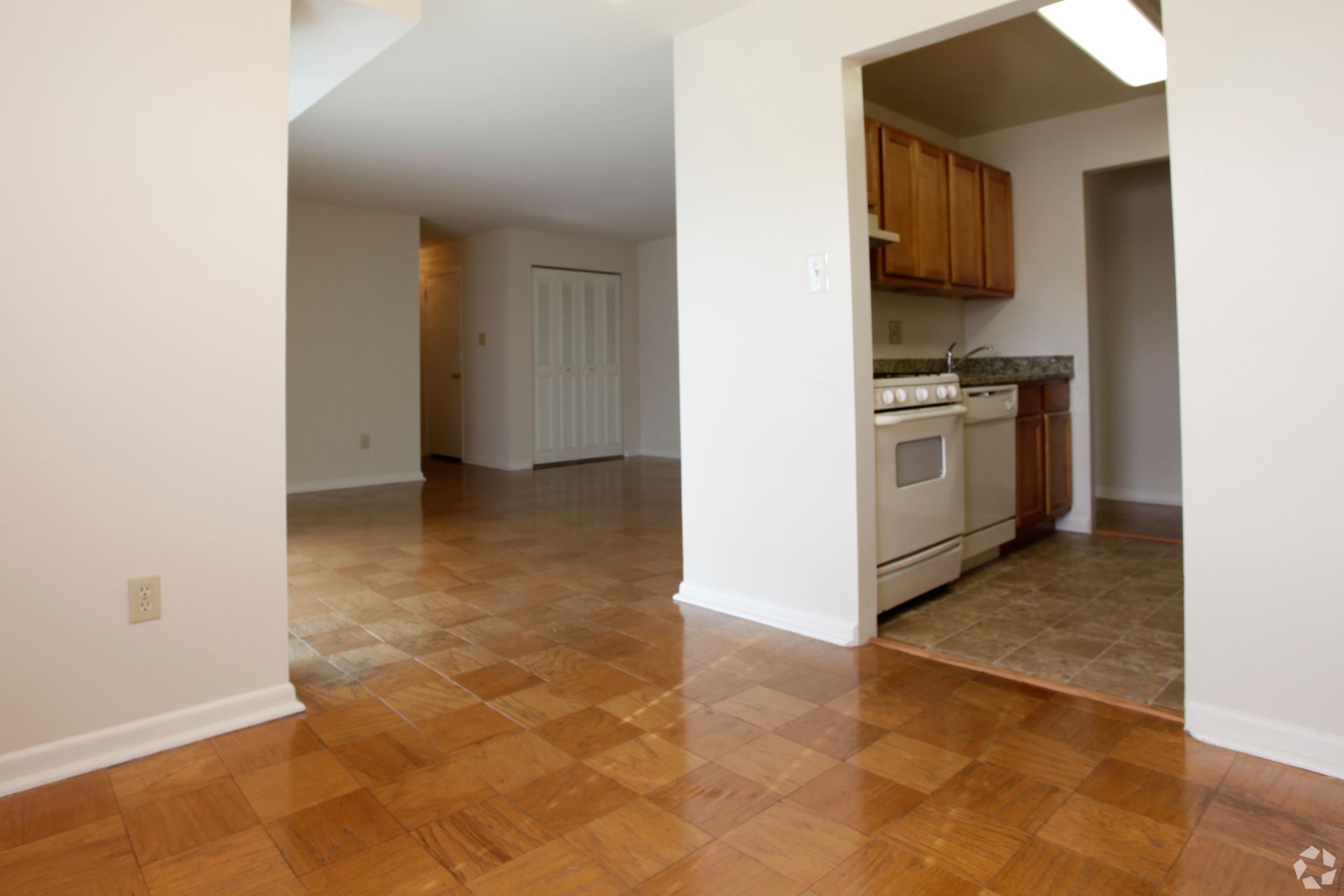 Interior of an apartment with hardwood floors, kitchen visible to the right, and a hallway to the left.