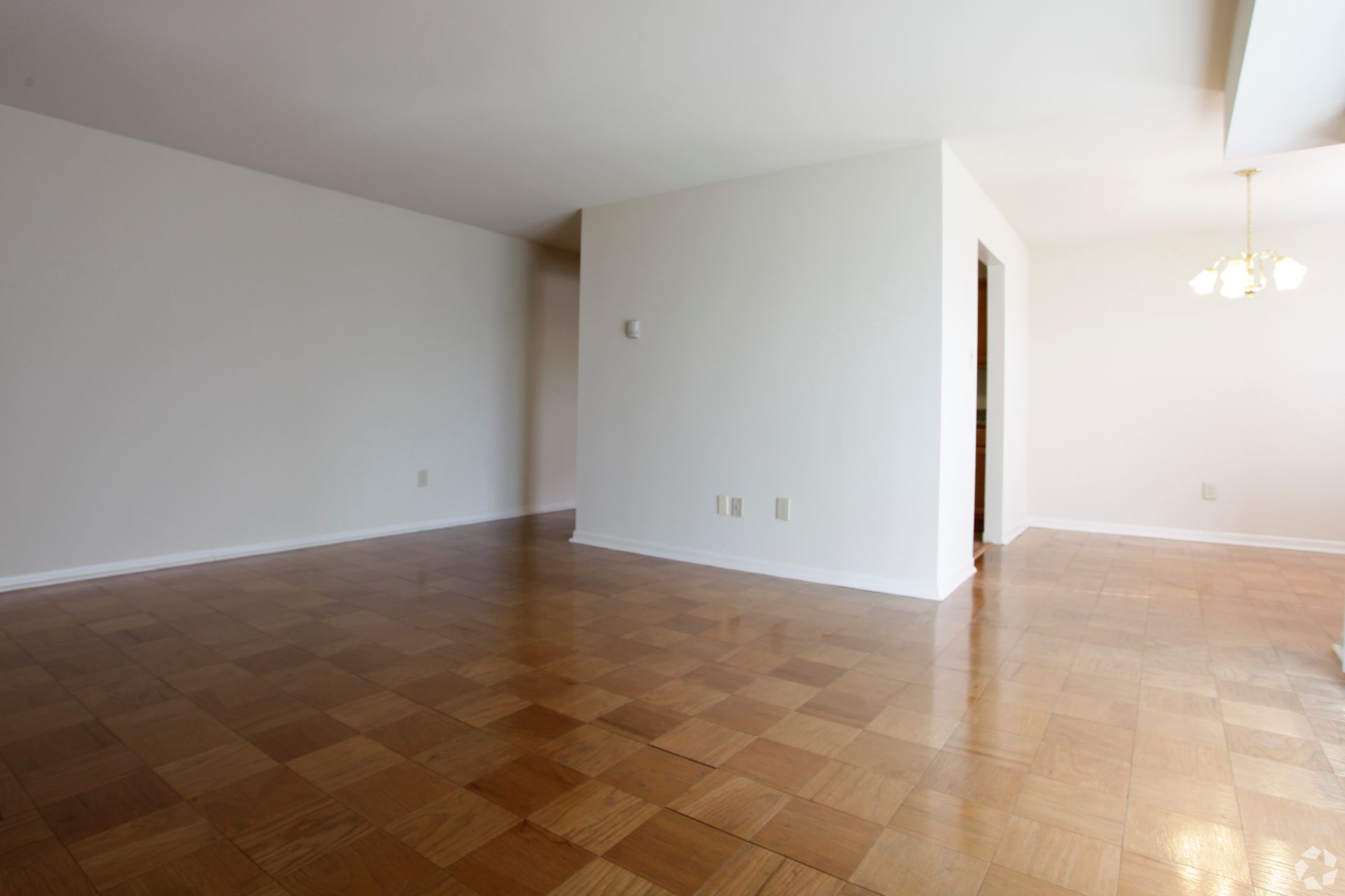 Empty room with wood floors and white walls. Doorway visible, chandelier.