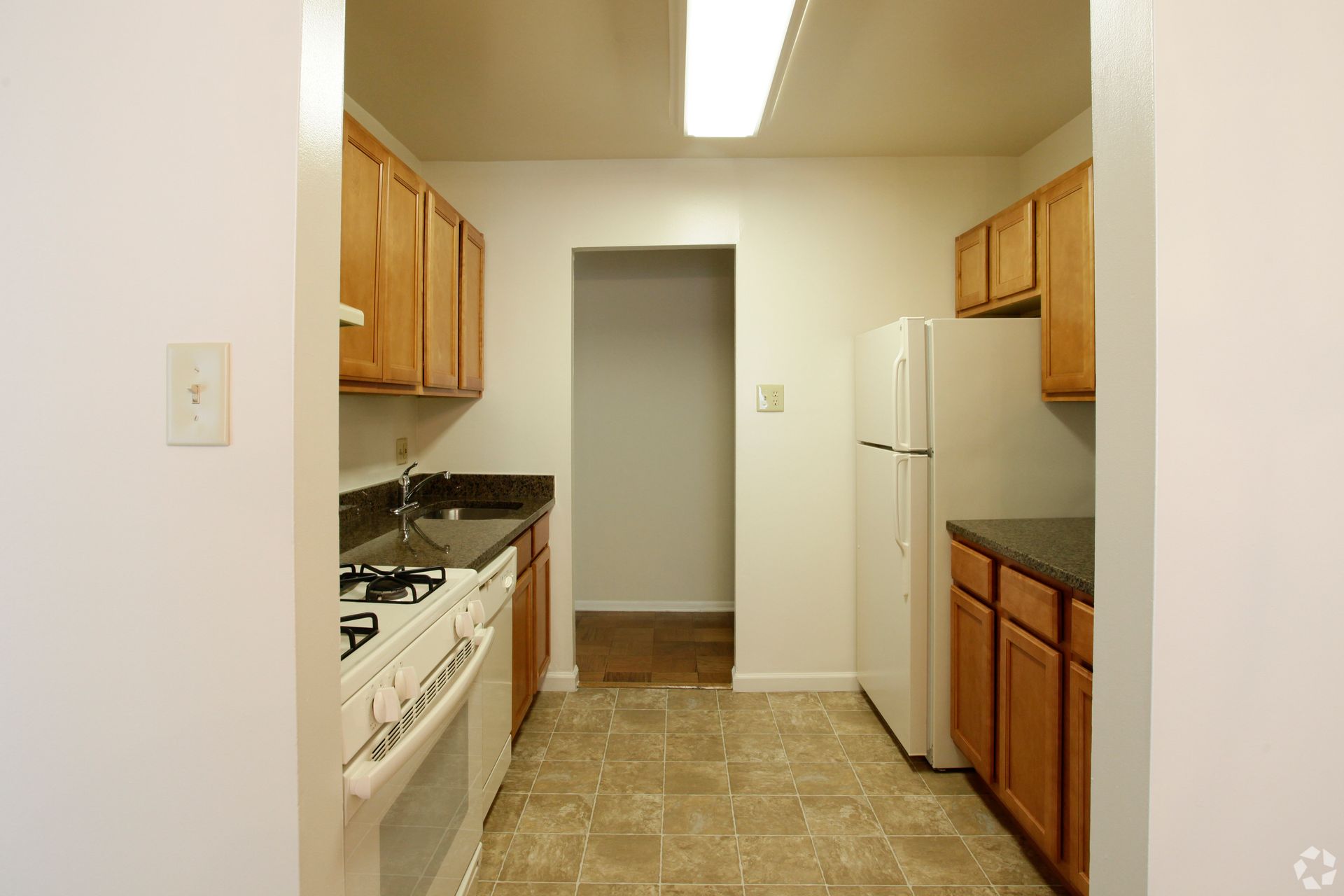 Small kitchen with wooden cabinets, white appliances, and a doorway at the end.