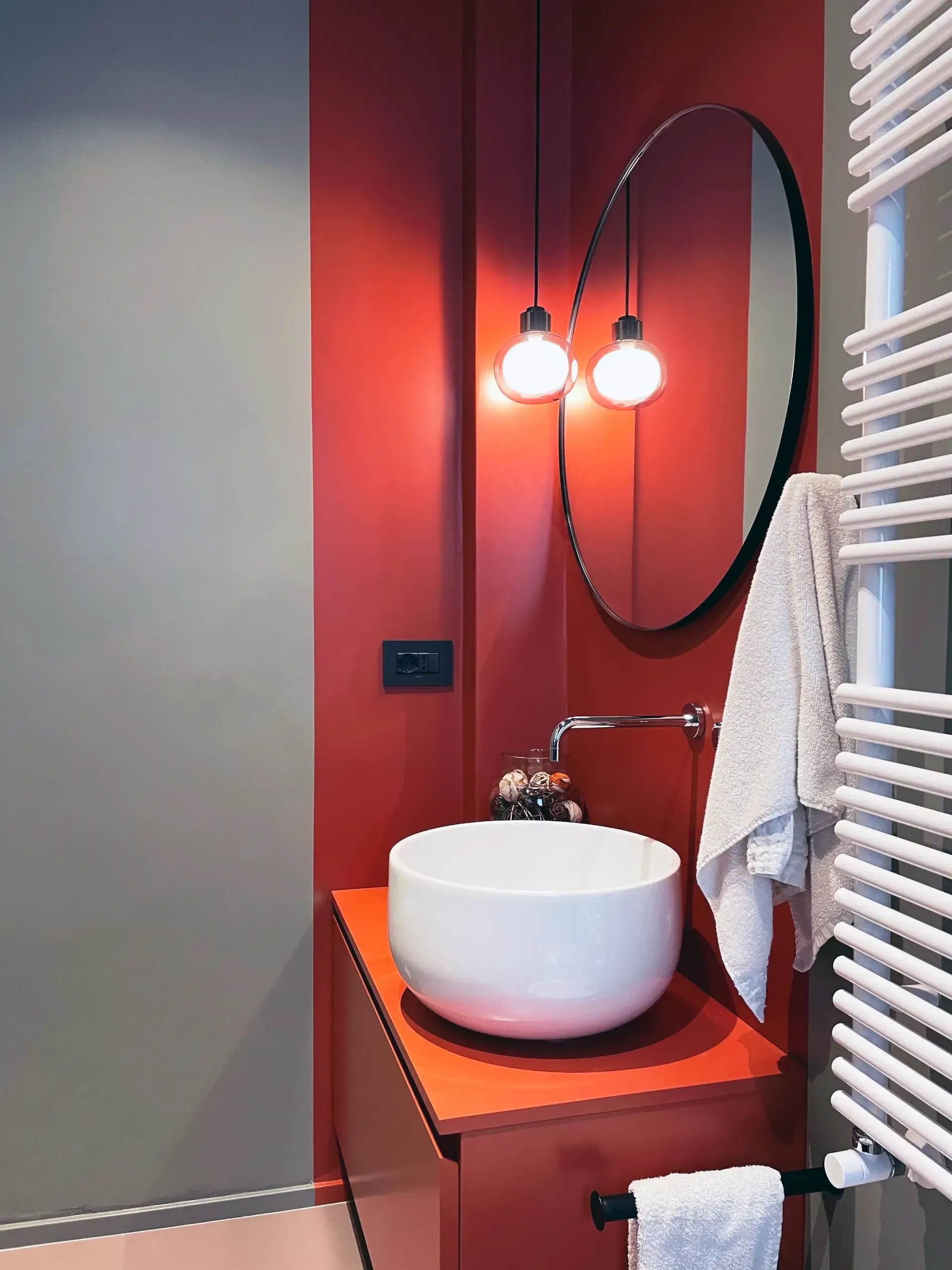 Bagno moderno con una combinazione di colori rosso e grigio, uno specchio rotondo e un lavandino bianco.