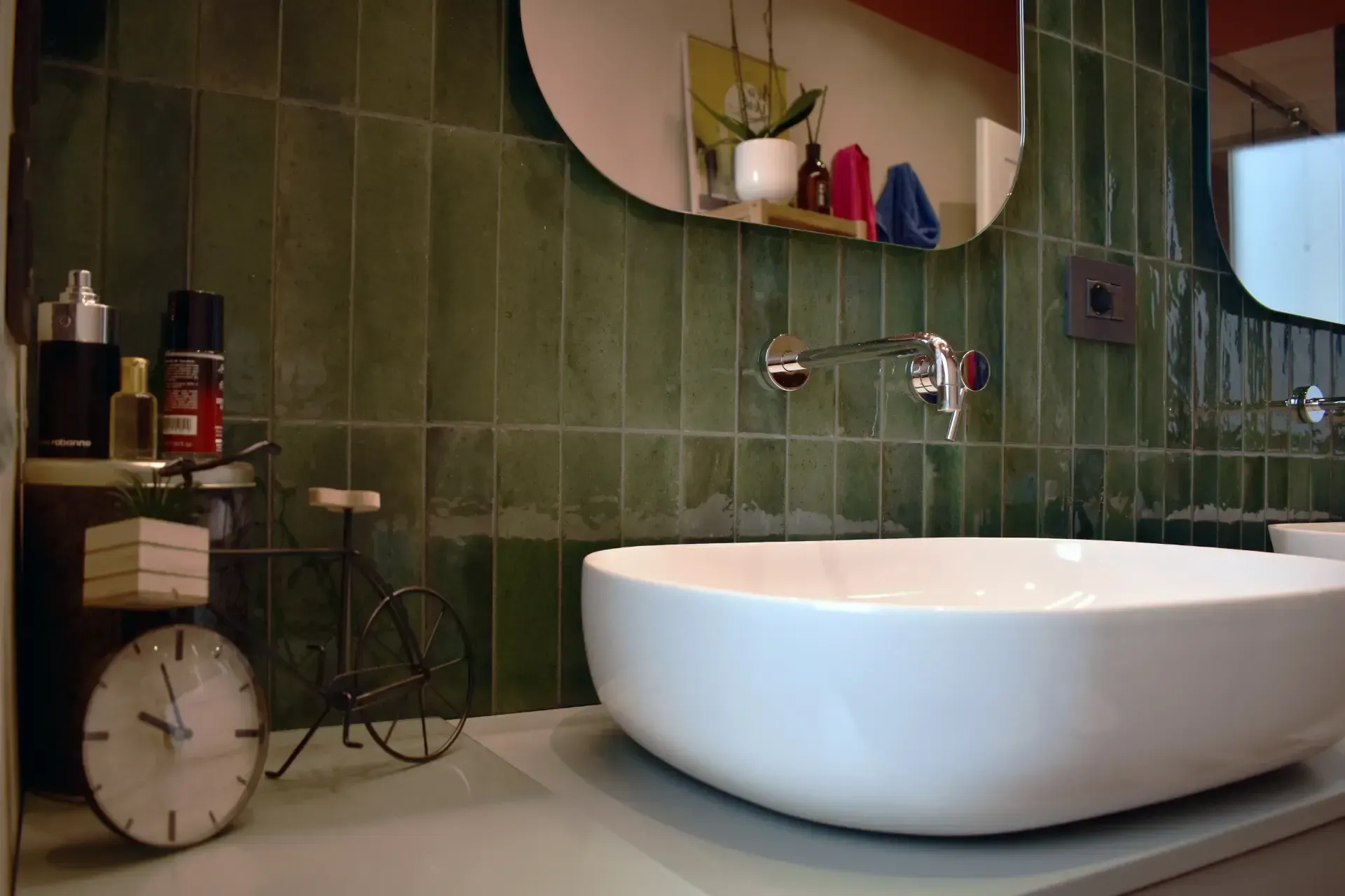 Lavandino bianco in un moderno bagno piastrellato di verde con specchio e articoli da toeletta.