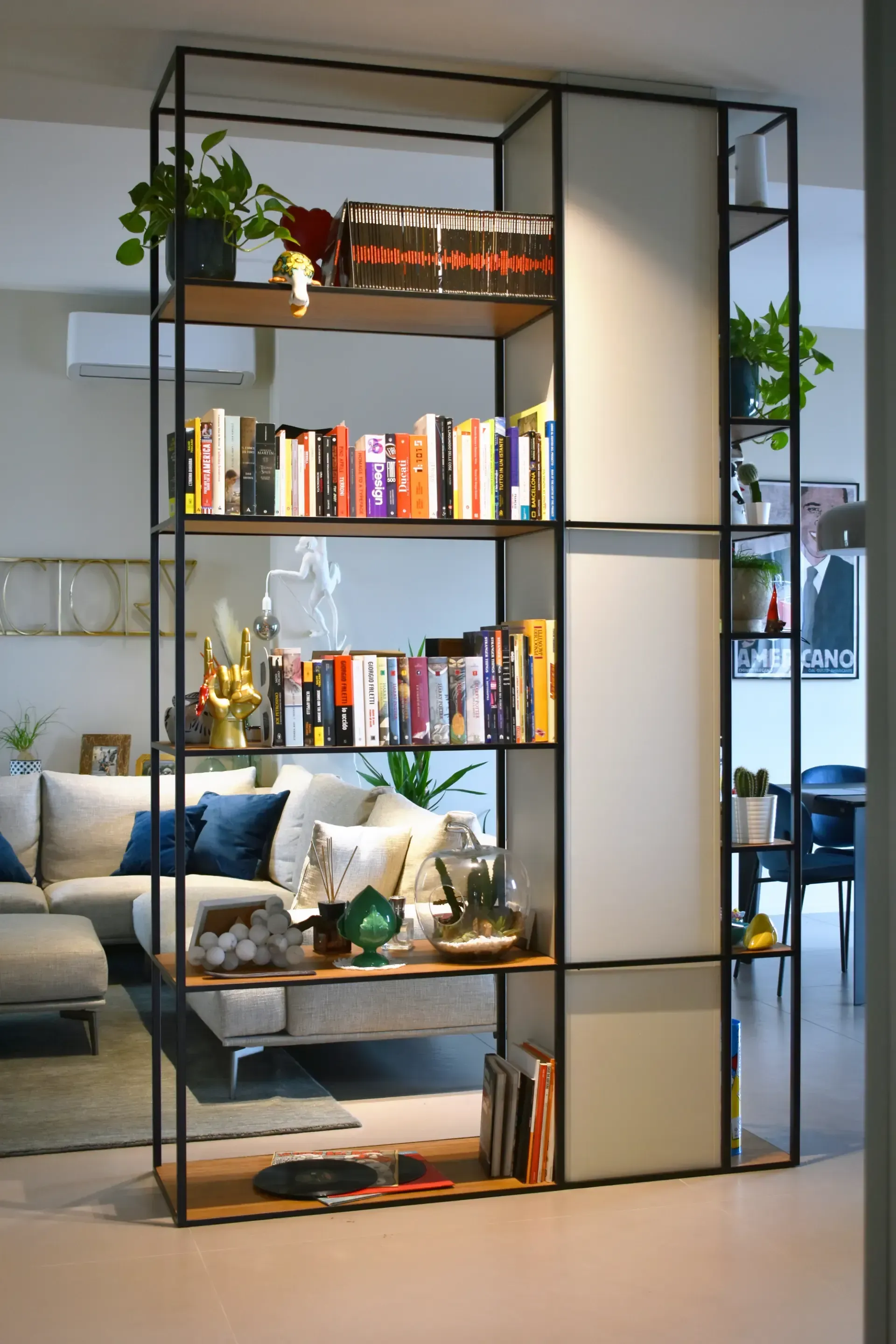 Libreria in metallo nero con libri, piante e oggetti decorativi, che divide lo spazio abitativo.
