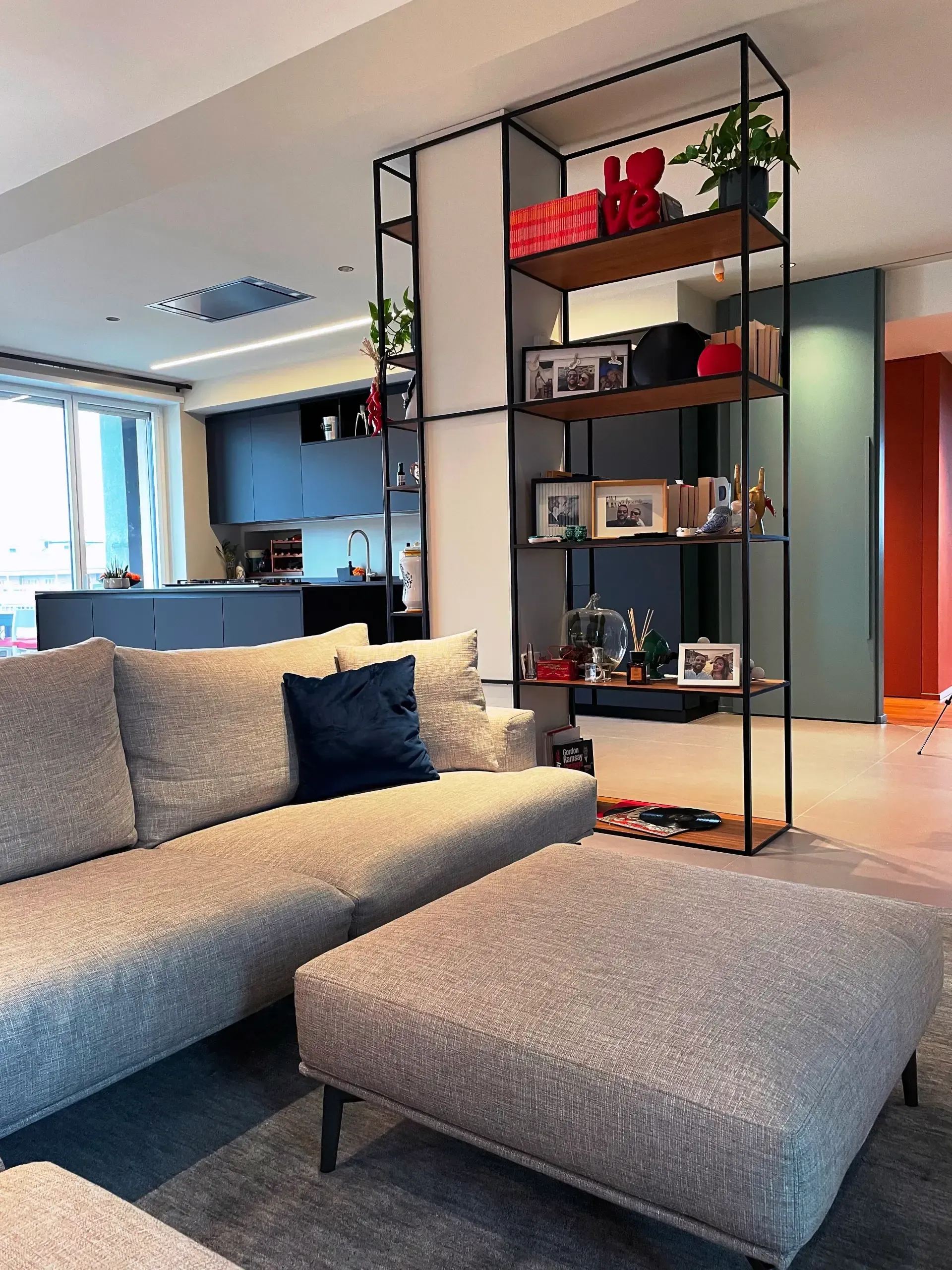 Soggiorno con divano grigio, pouf, scaffalatura nera e cucina a vista.