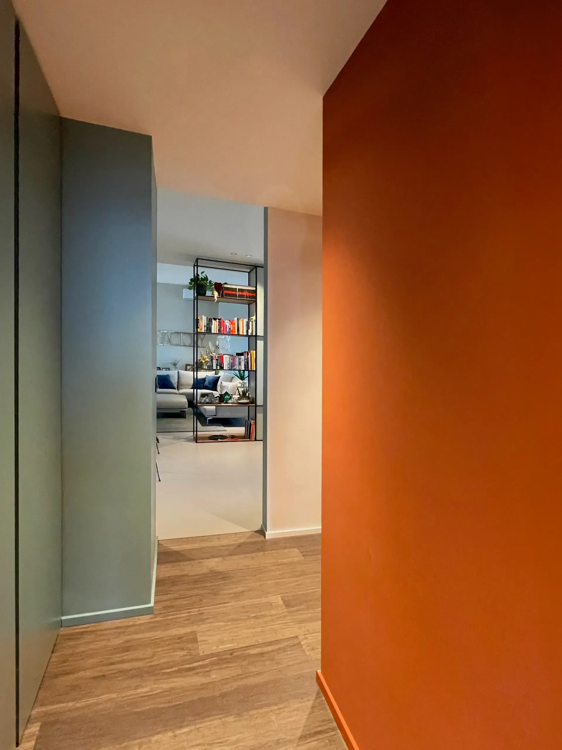 Corridoio con pareti arancioni e verdi, che conduce a una stanza con scaffali e mobili; pavimento in legno.