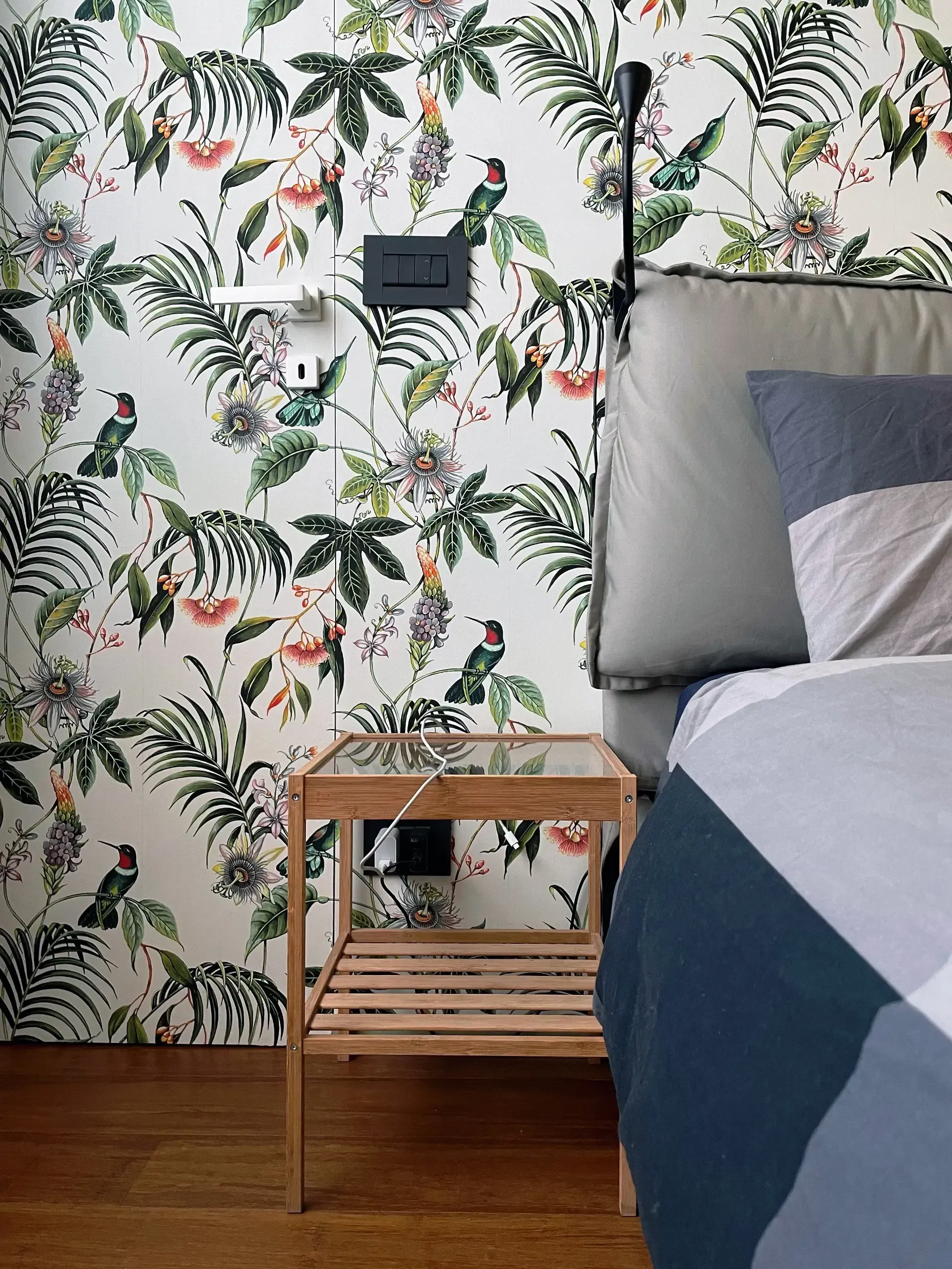 Camera da letto con carta da parati floreale, comodino in legno e letto con cuscini grigi e lenzuola blu.