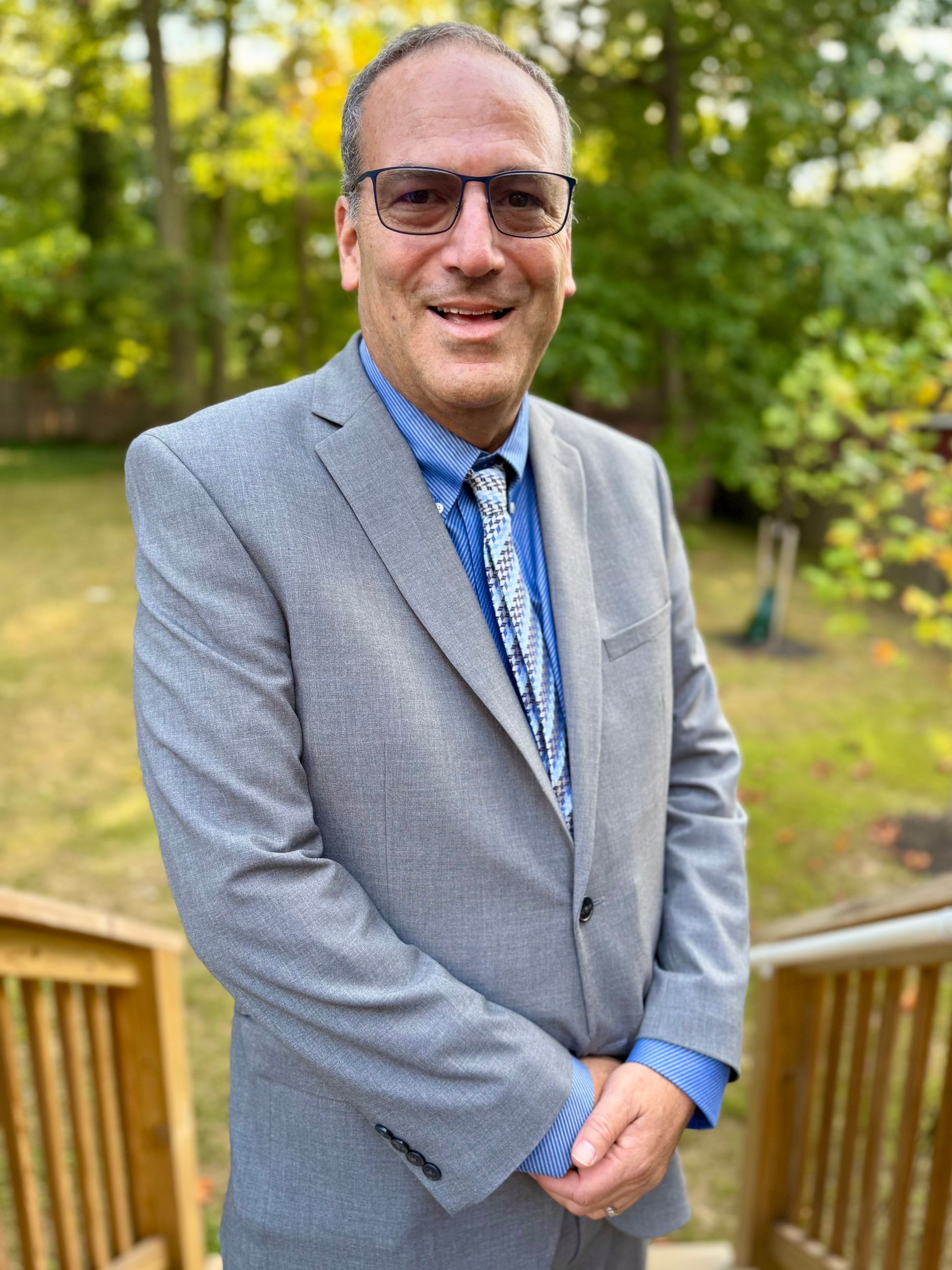 Man in a blue suit and colorful tie, smiling, standing in front of a blue background.