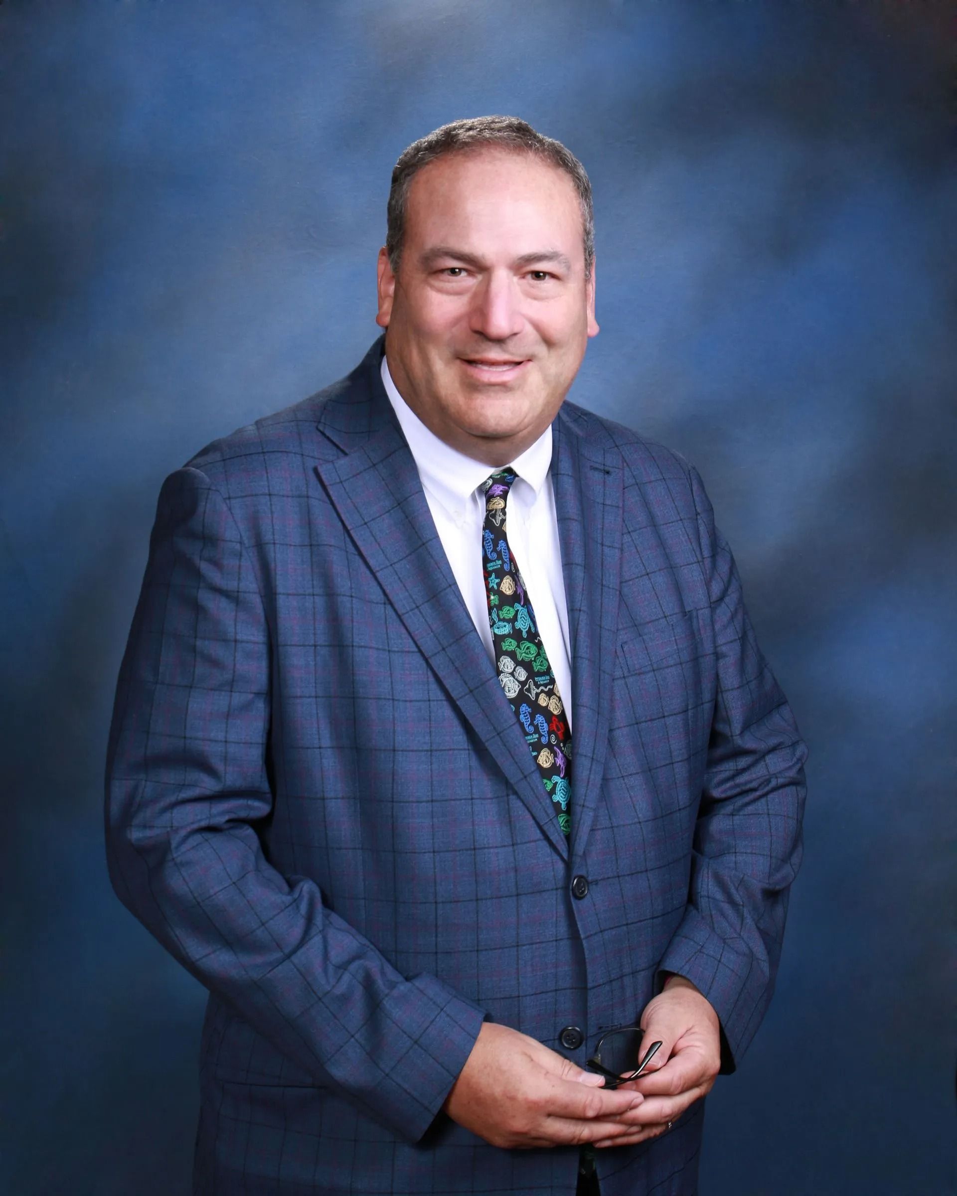 Man in a blue suit and colorful tie, smiling, standing in front of a blue background.