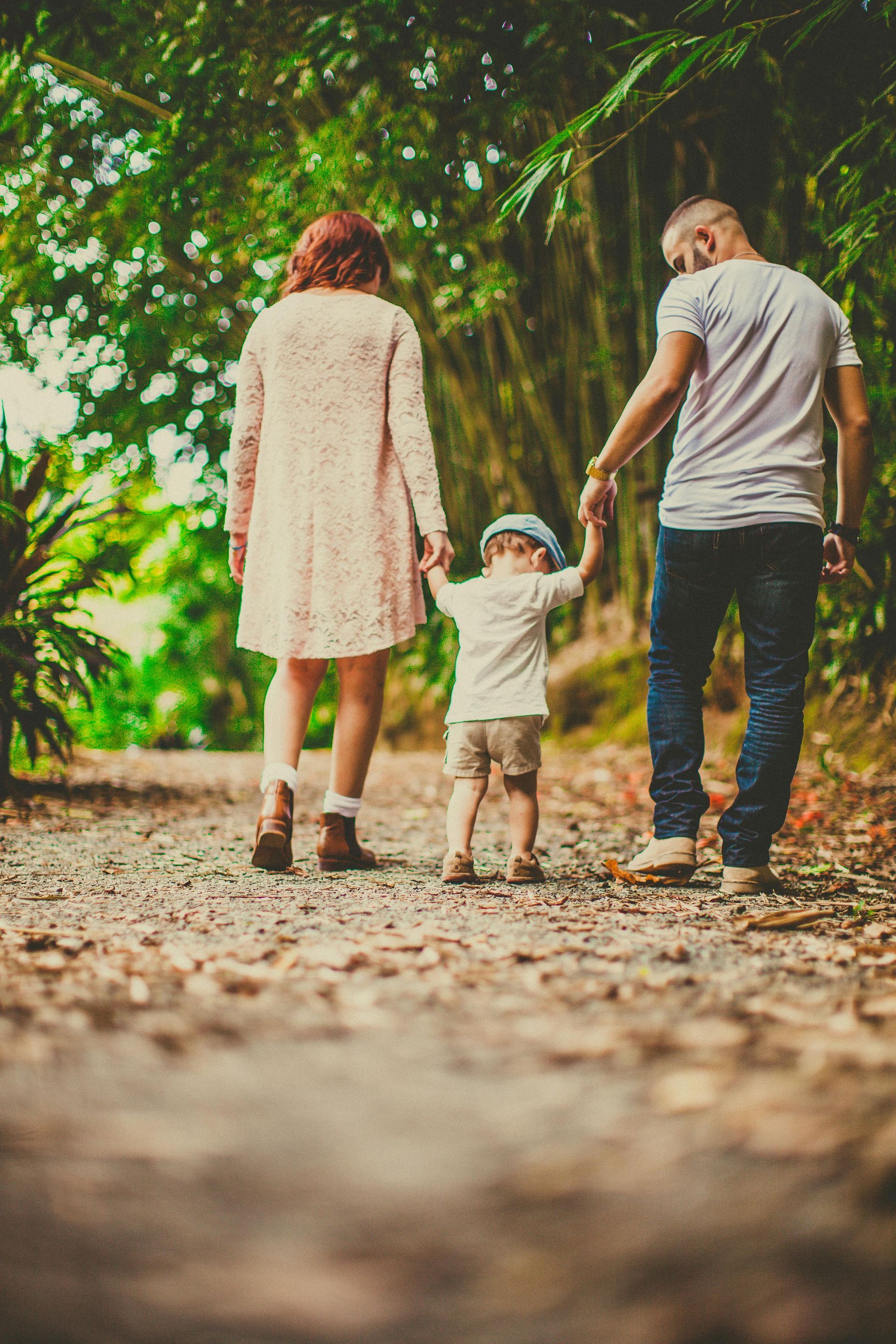 Family of three walking away on a wooded path. A woman in a pink dress and a man in jeans hold hands with a small child.