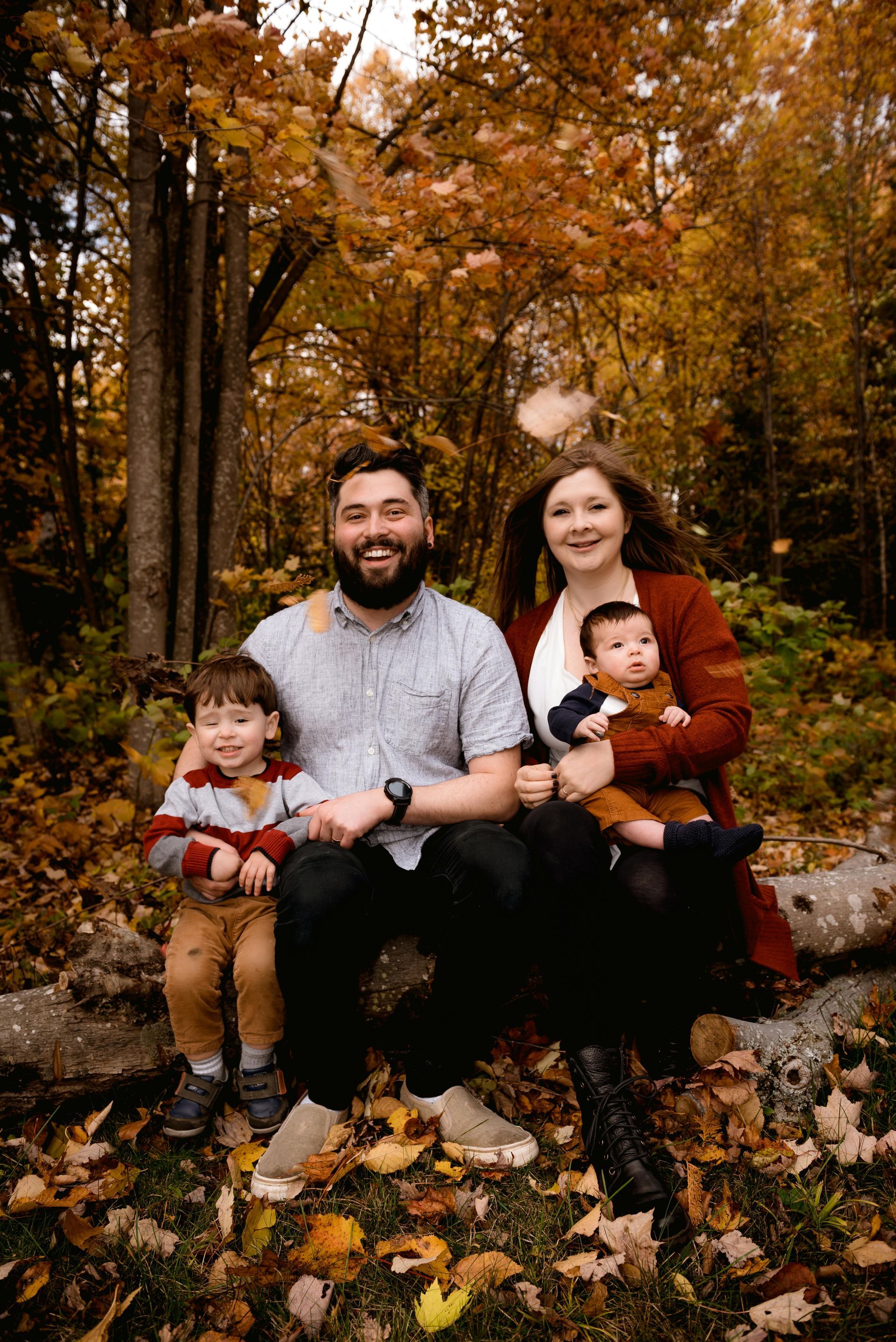 Family of four poses on a log in a forest with autumn leaves. The parents hold their children, everyone smiling.