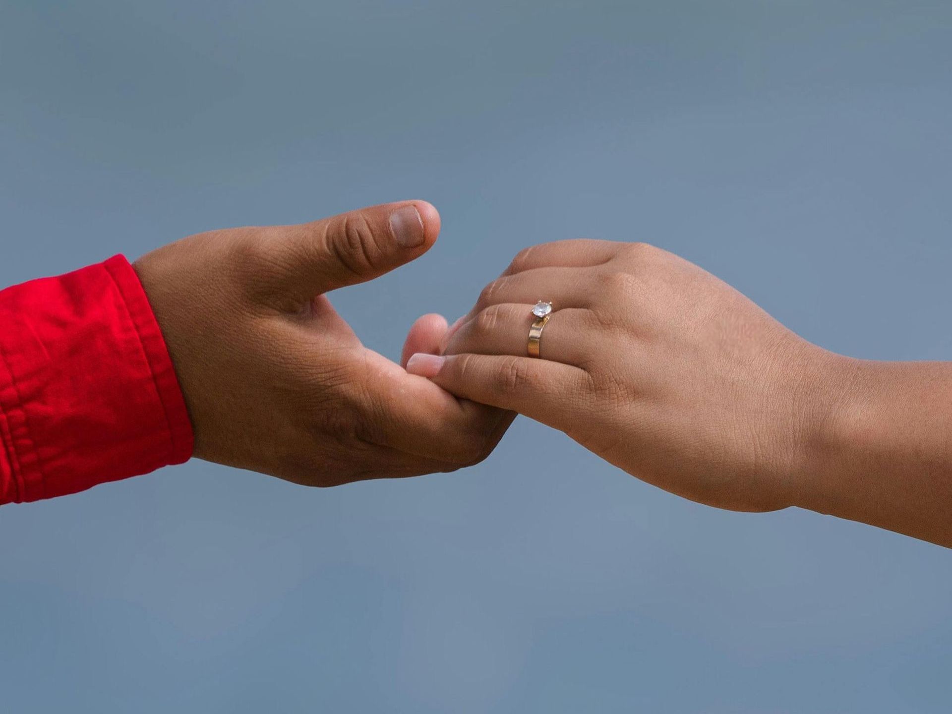 Two hands, one wearing a ring, gently touching against a blue background. The left sleeve is red.