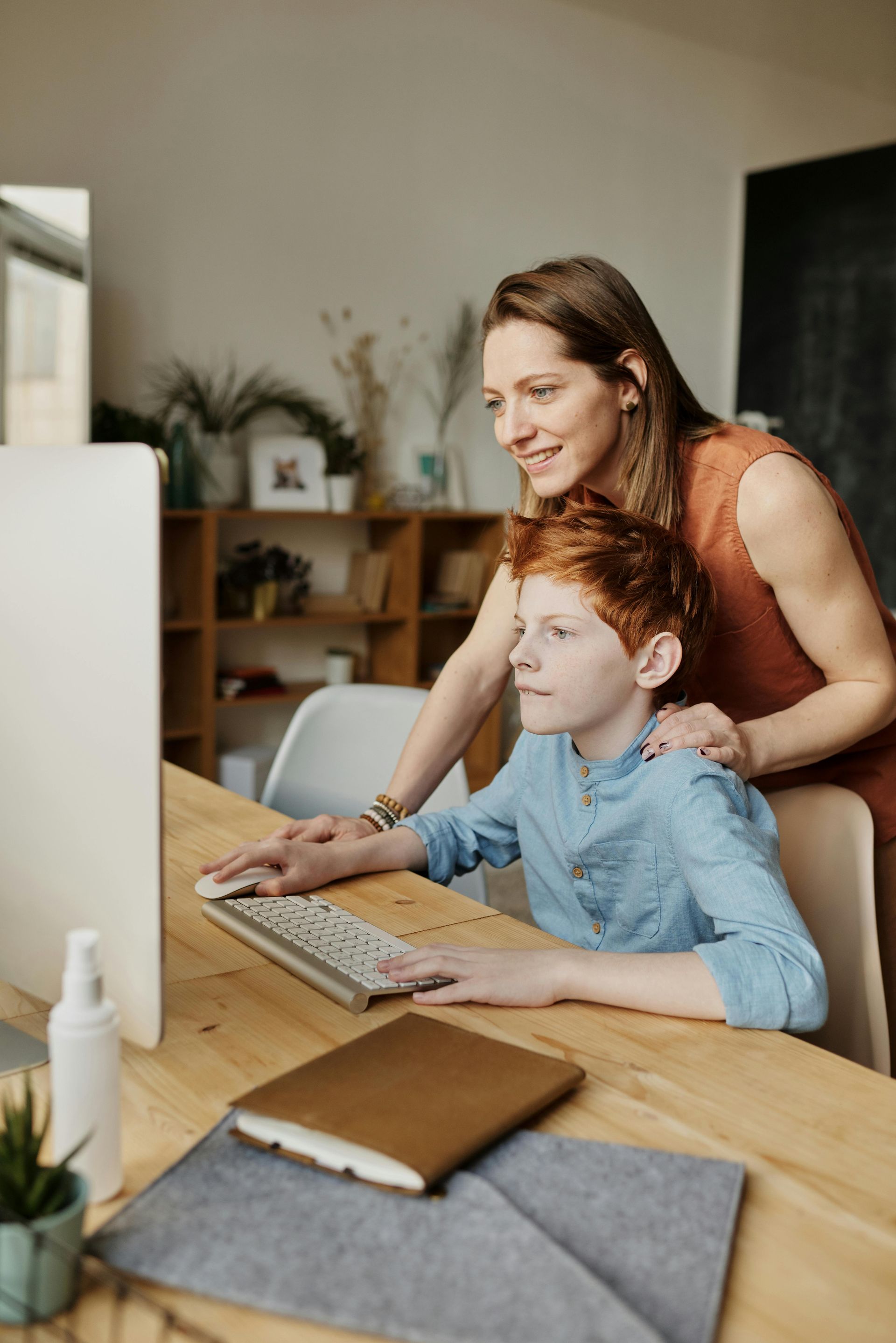 Woman assists a red-haired boy at a computer. Both are looking at the screen, woman with a smile, in a room with wooden furniture.