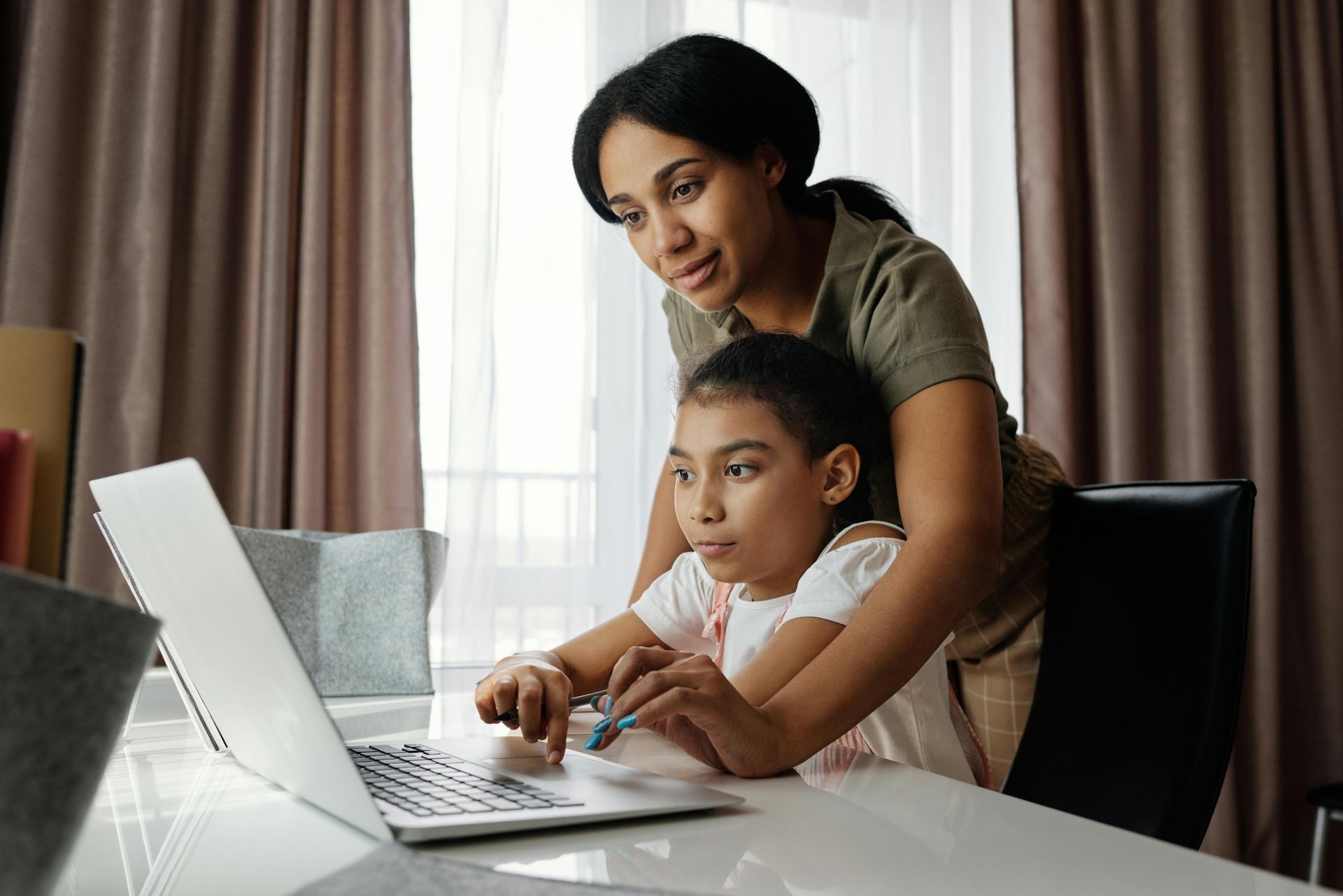 A woman guides a young girl's hands on a laptop at a table near a window. Both are looking at the screen.