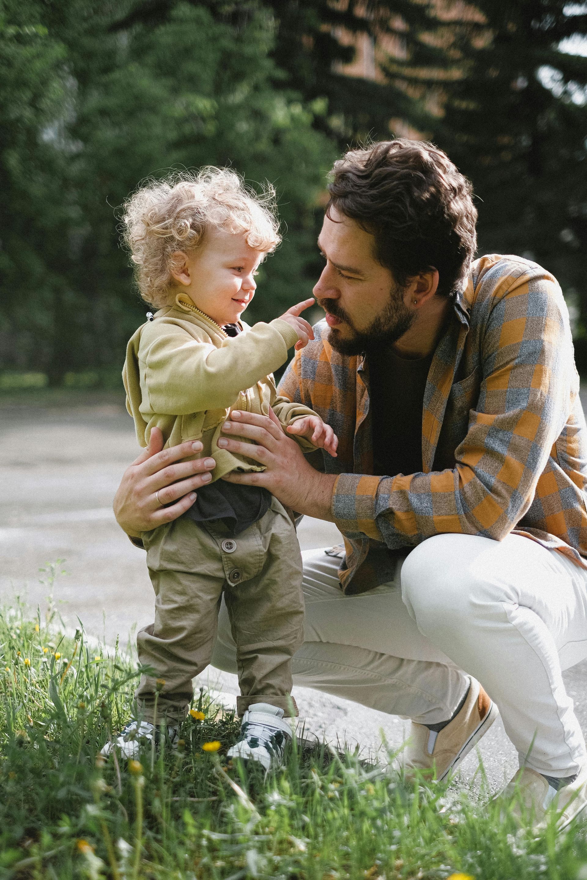 A child with curly blonde hair playfully pokes the nose of a bearded man squatting outdoors in a park-like setting.