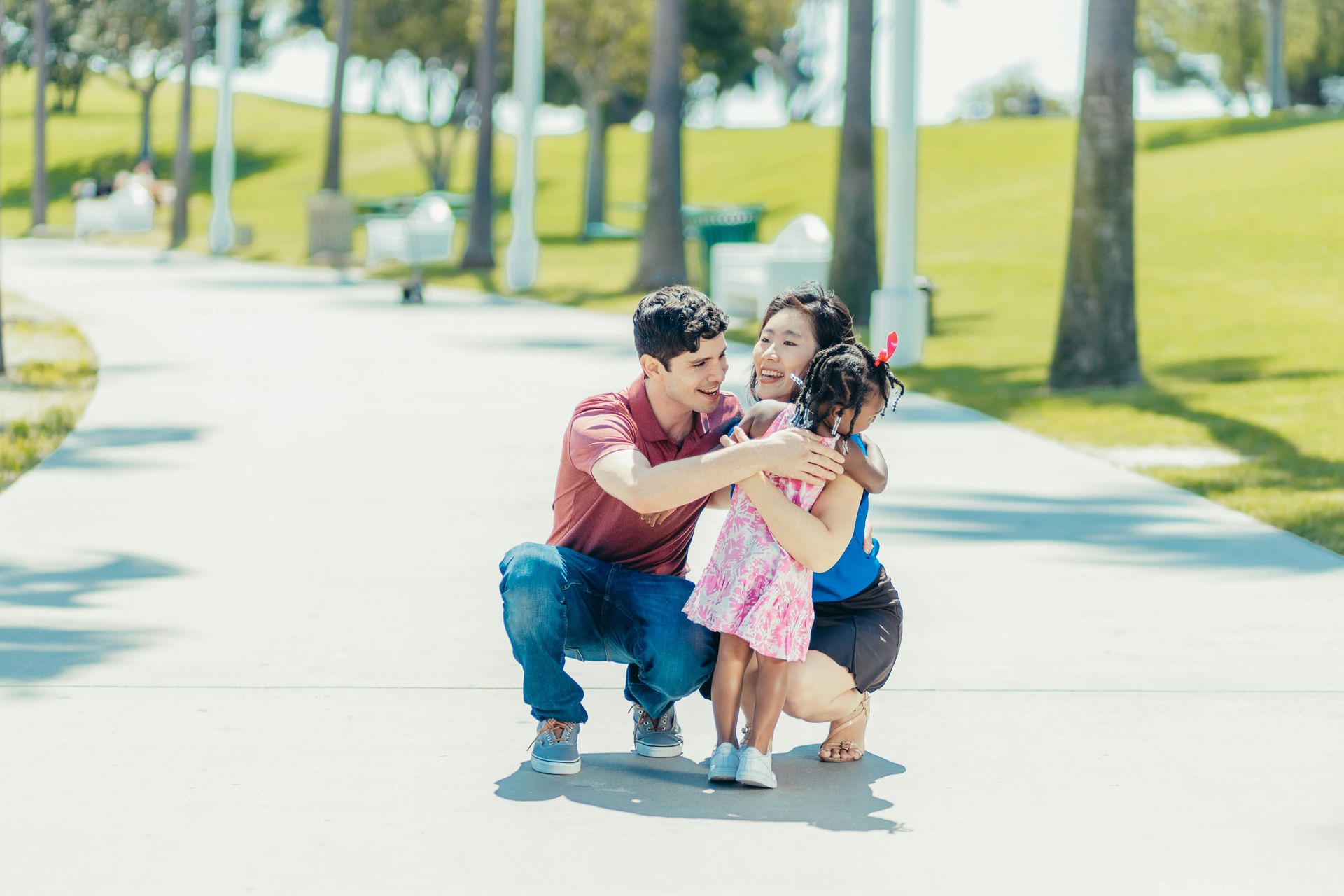 A family of three embraces on a sunny path in a park. The parents, a man and woman, are kneeling to hug their young child.