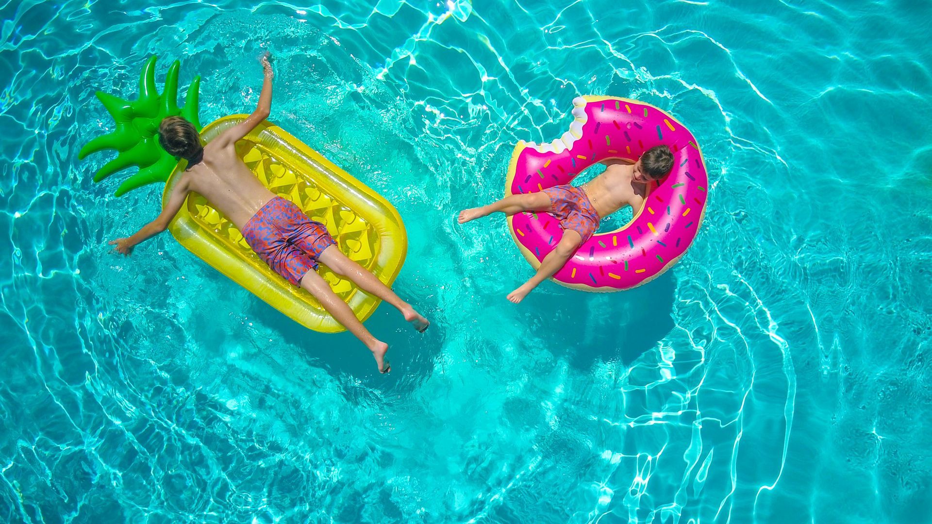 Two people in a pool on inflatable floats: a pineapple and a pink donut.