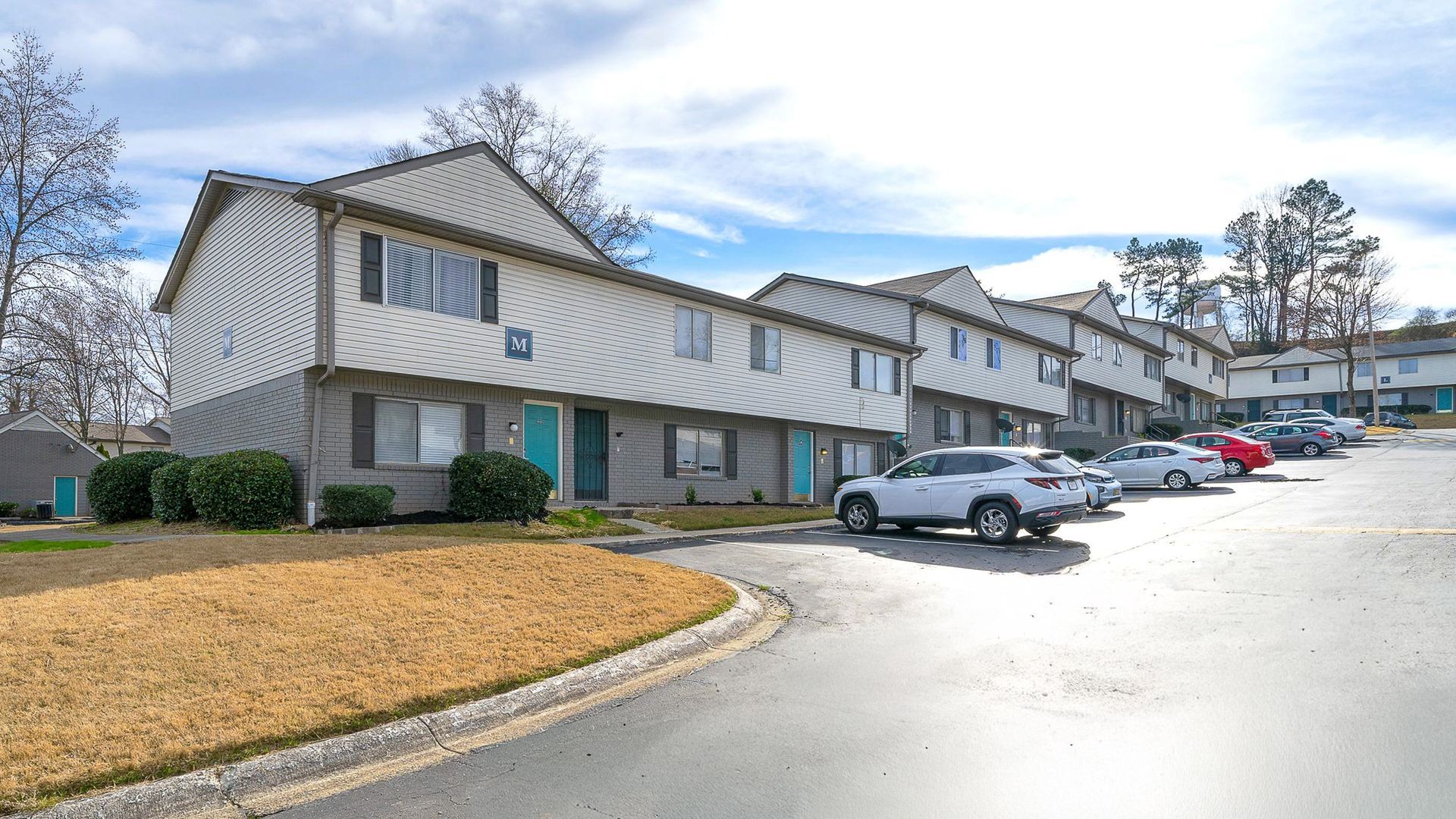 Row of two-story townhouses with turquoise doors, parked cars on the side, and grassy area.
