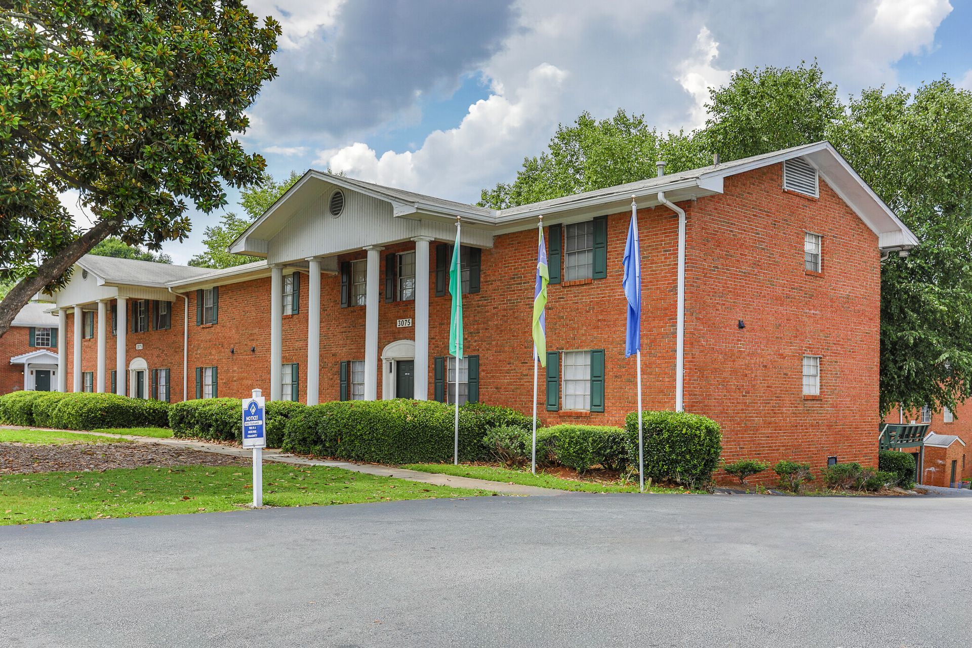 Red brick apartment building with white columns and flags; green bushes and trees.