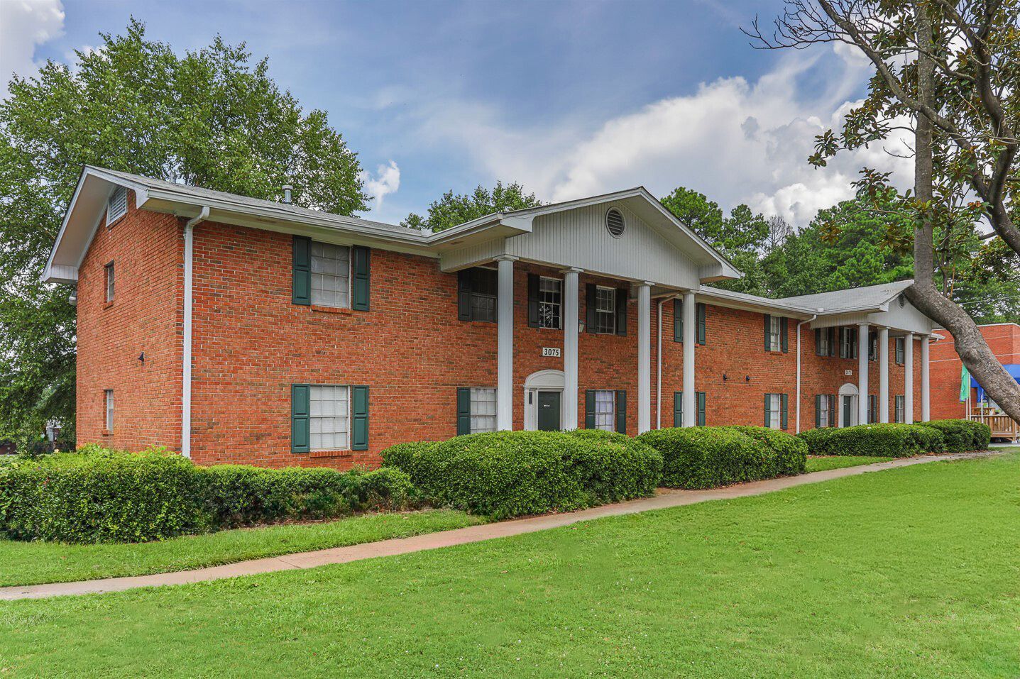 Red brick apartment building with white columns and green shutters, set in a grassy yard under a cloudy sky.