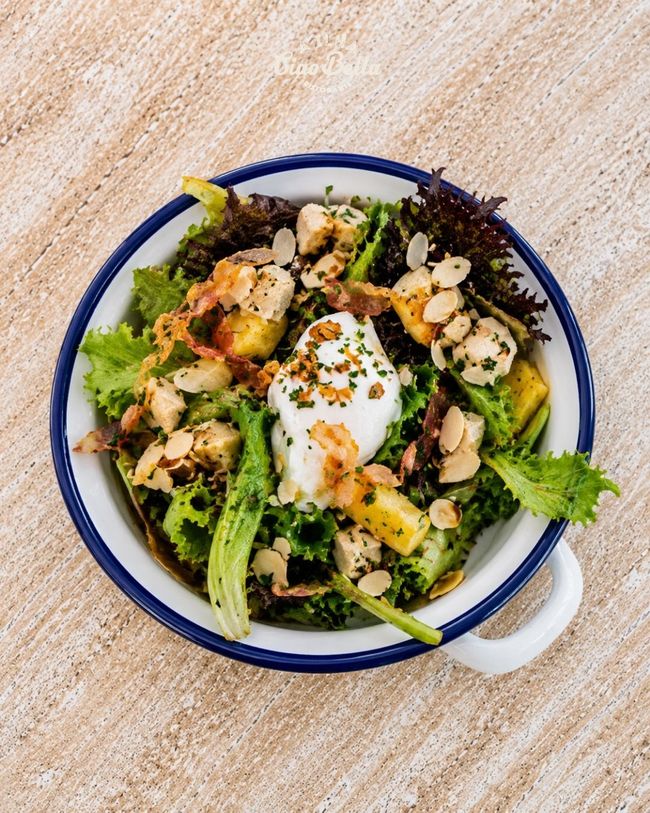 Salada de legumes grelhados com ovo mole em uma tigela com borda azul sobre uma mesa de madeira clara.
