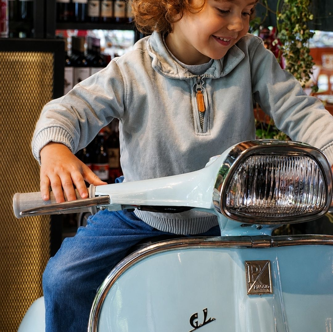 Criança sorridente sentada em uma scooter azul-clara, segurando o guidão em um café.