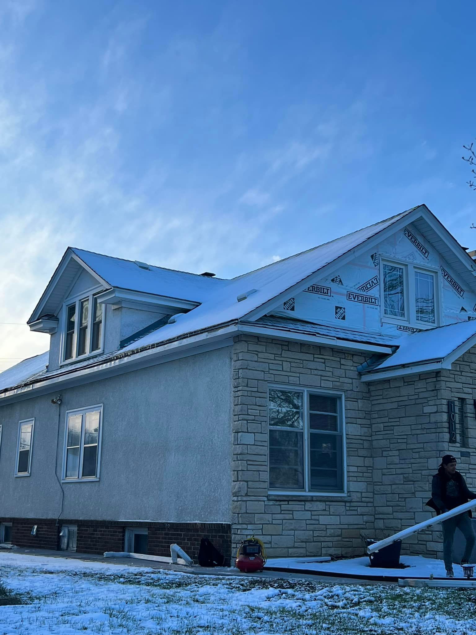 A man is standing in front of a house with snow on the roof.