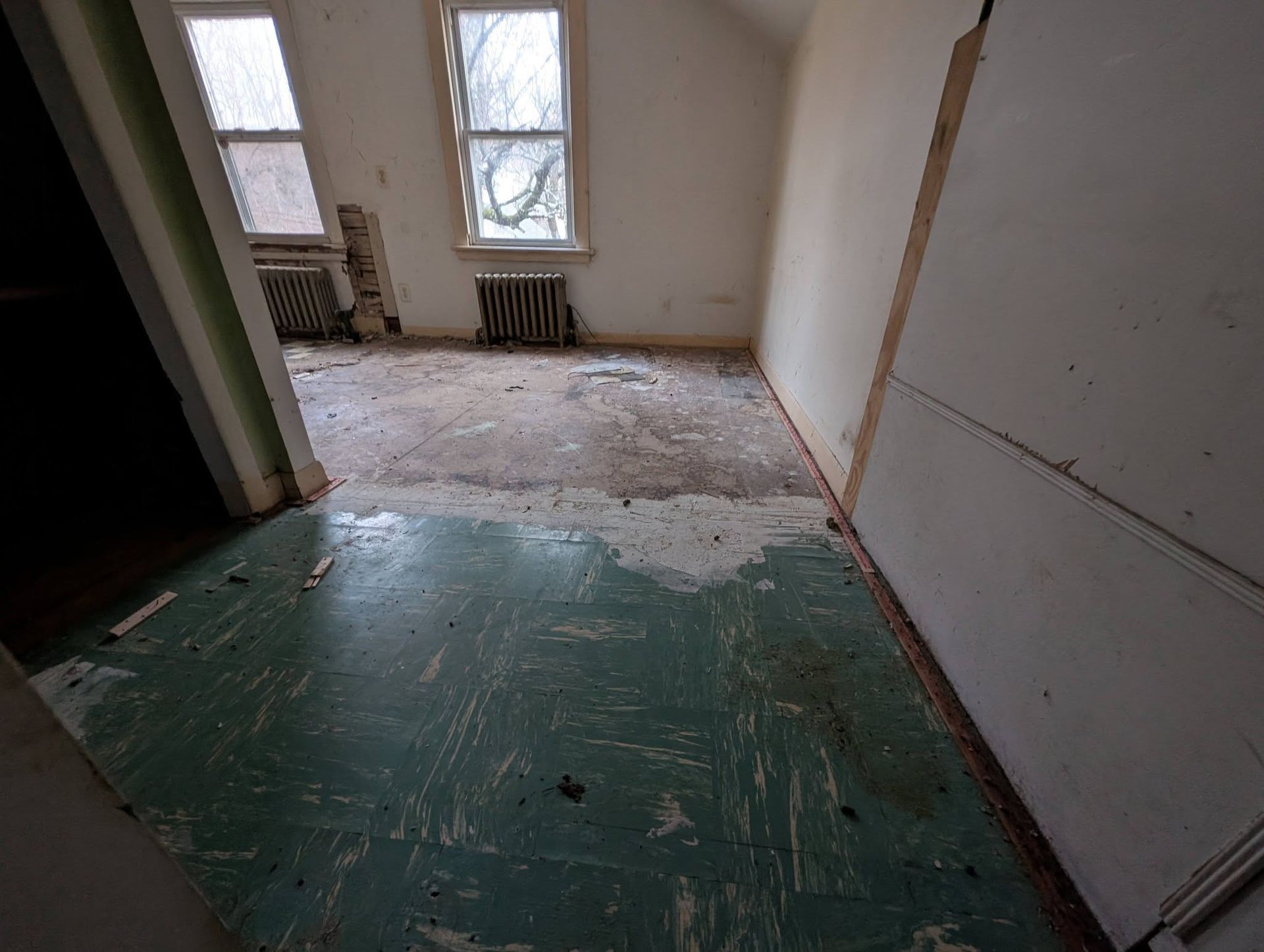 Dilapidated room with peeling green floor, two windows, and exposed wall.
