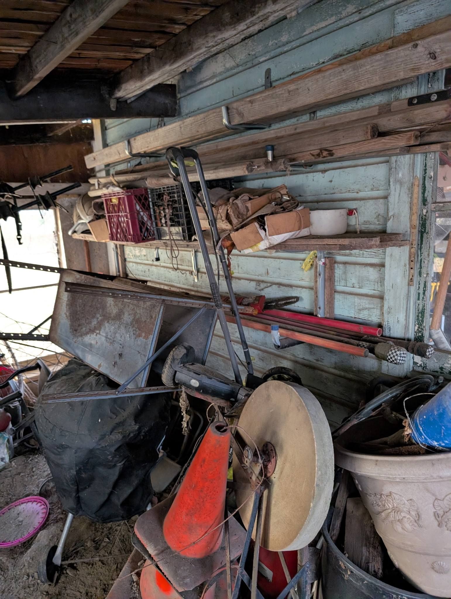 Old workshop with tools and supplies. A grinder, lawnmower, and various items sit under wooden shelving.