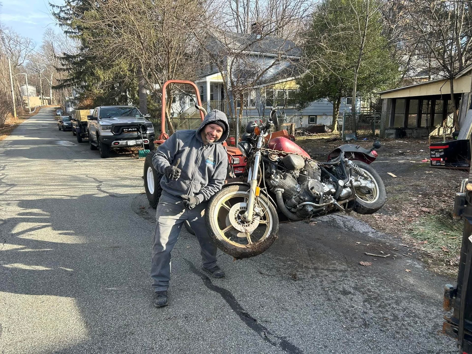 Man smiling, holding front wheel of fallen motorcycle on a paved road next to a tractor and truck.