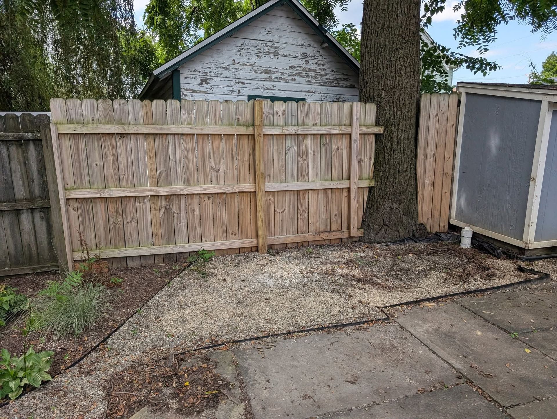 A weathered wooden fence surrounds a gravel patch, tree trunk, and an enclosed wooden box.