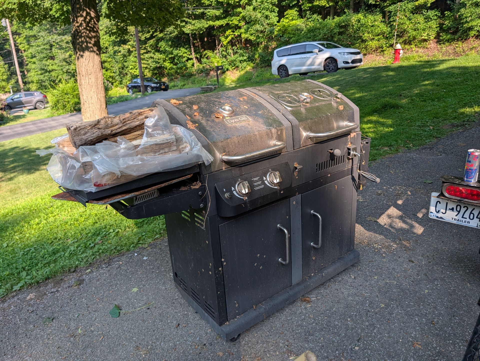 Black outdoor grill with three burners, surrounded by trees and parked cars.