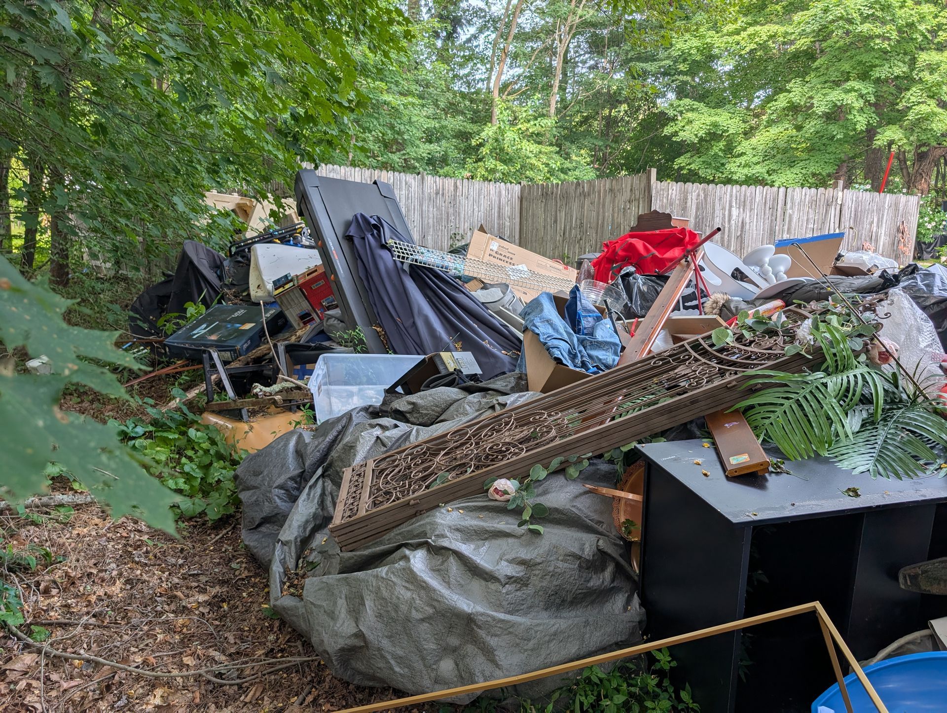 Pile of assorted trash in a backyard, including furniture, plastic bags, and boxes, near a wooden fence.